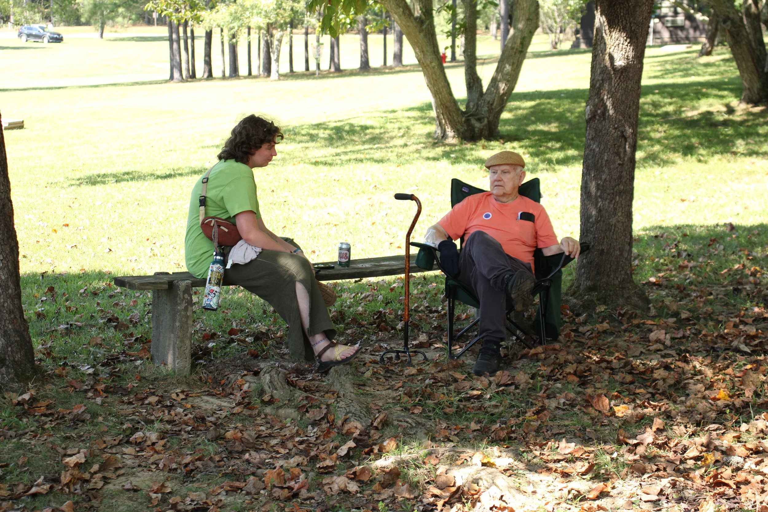 A young woman and an elderly man are sitting under a tree in a park. The woman, wearing a green shirt and brown pants, is sitting on a wooden bench, while the man, wearing an orange shirt, black pants, and a tan cap, is seated on a portable chair with a walking cane between them. They are having a conversation, with the woman holding a little object and a drink can on a nearby table.