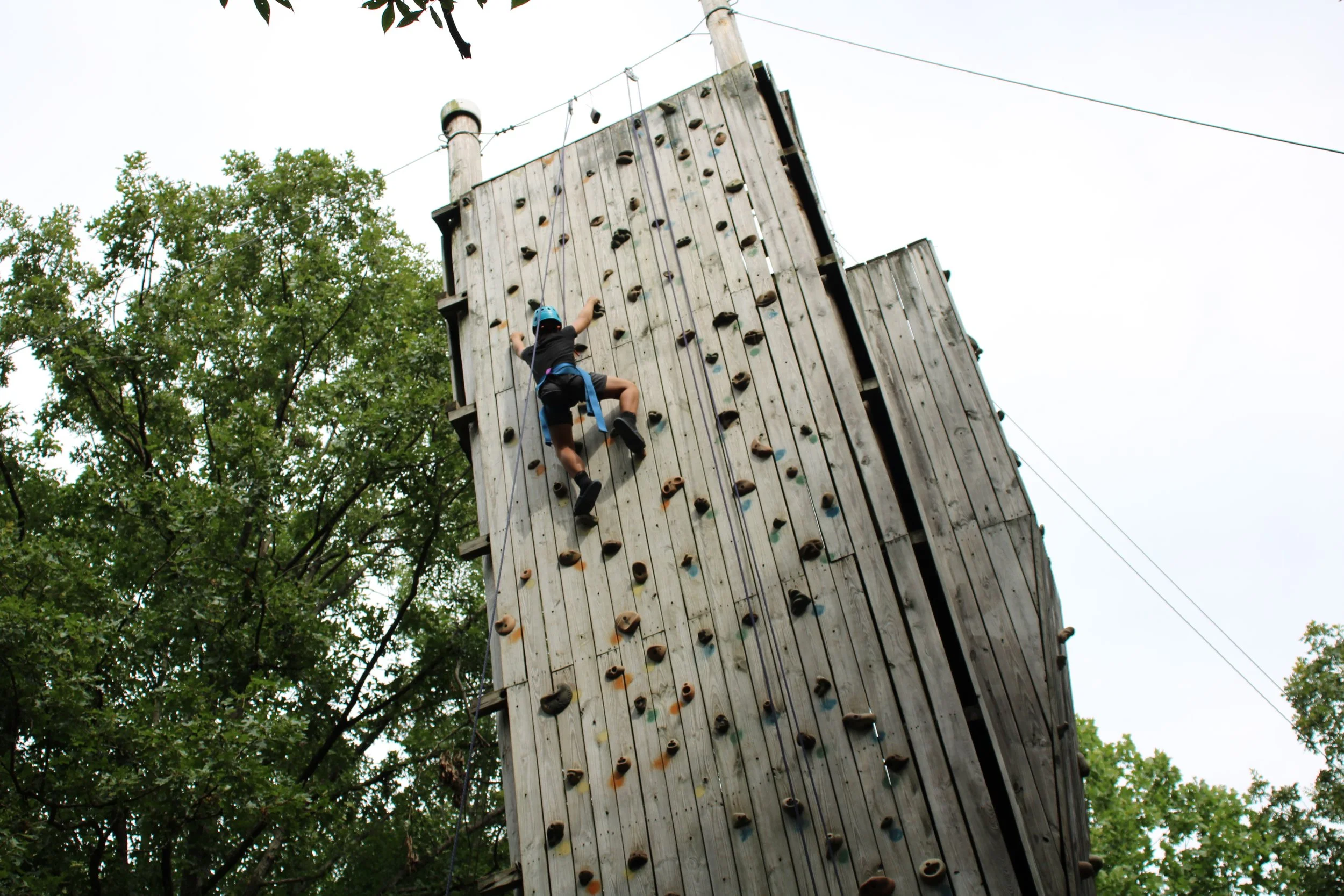 Young boy wearing a safety helmet and harness climbing an outdoor wooden rock wall with colorful handholds, surrounded by green trees.