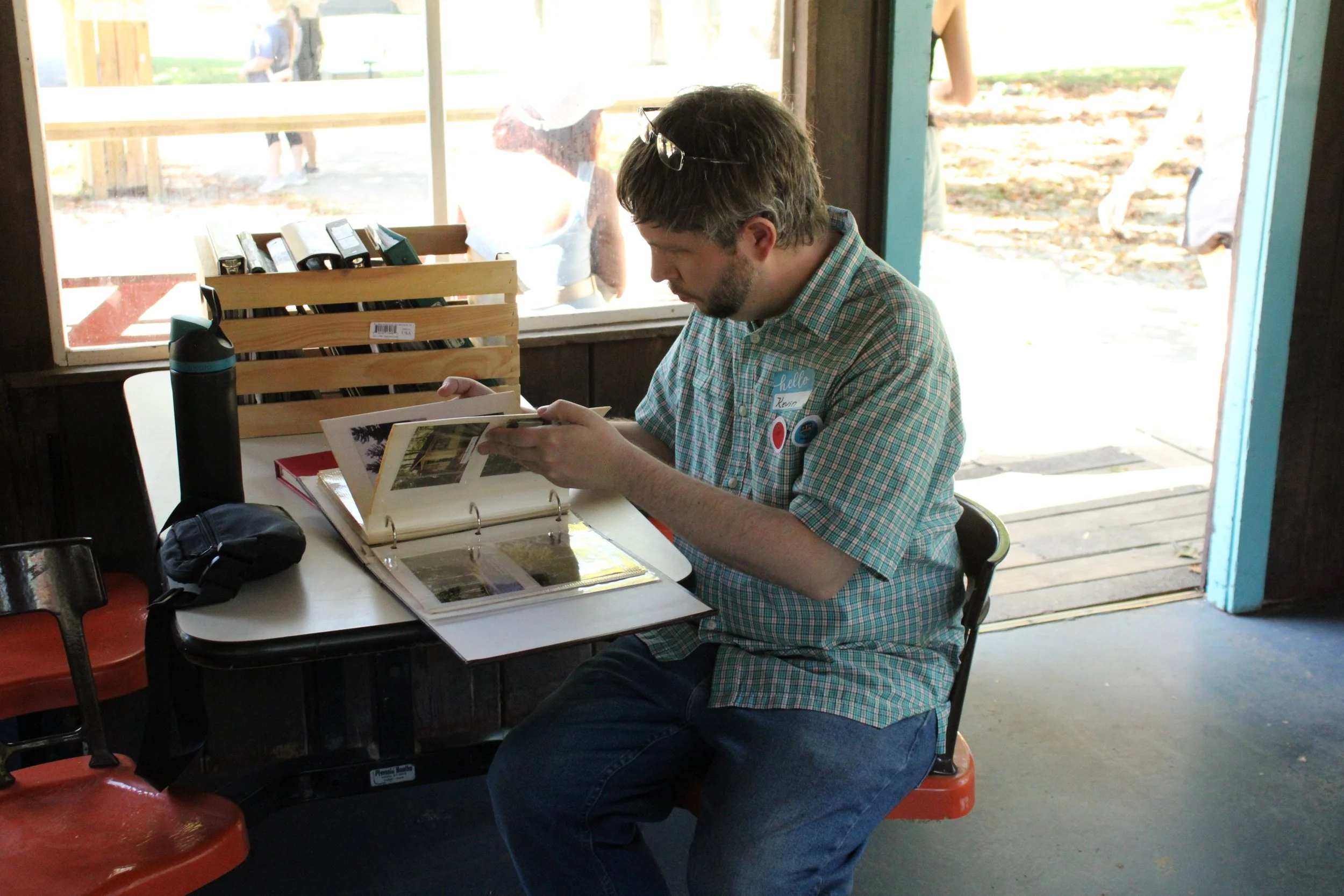 A man sitting at a table inside, looking at photo albums, with a water bottle and black bag on the table, a window behind him revealing outdoor scenery.
