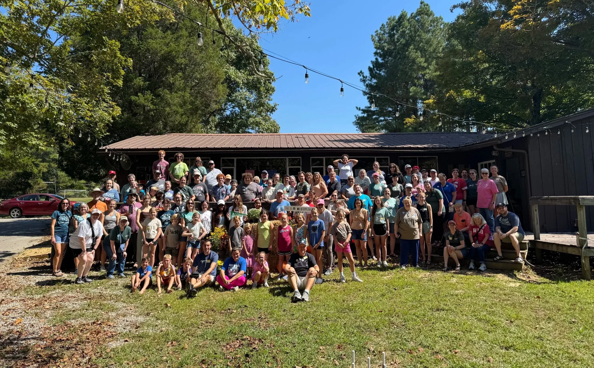 A large group of people gathered outside in front of a black building, under string lights, with trees and a blue sky in the background, posing for a group photo.