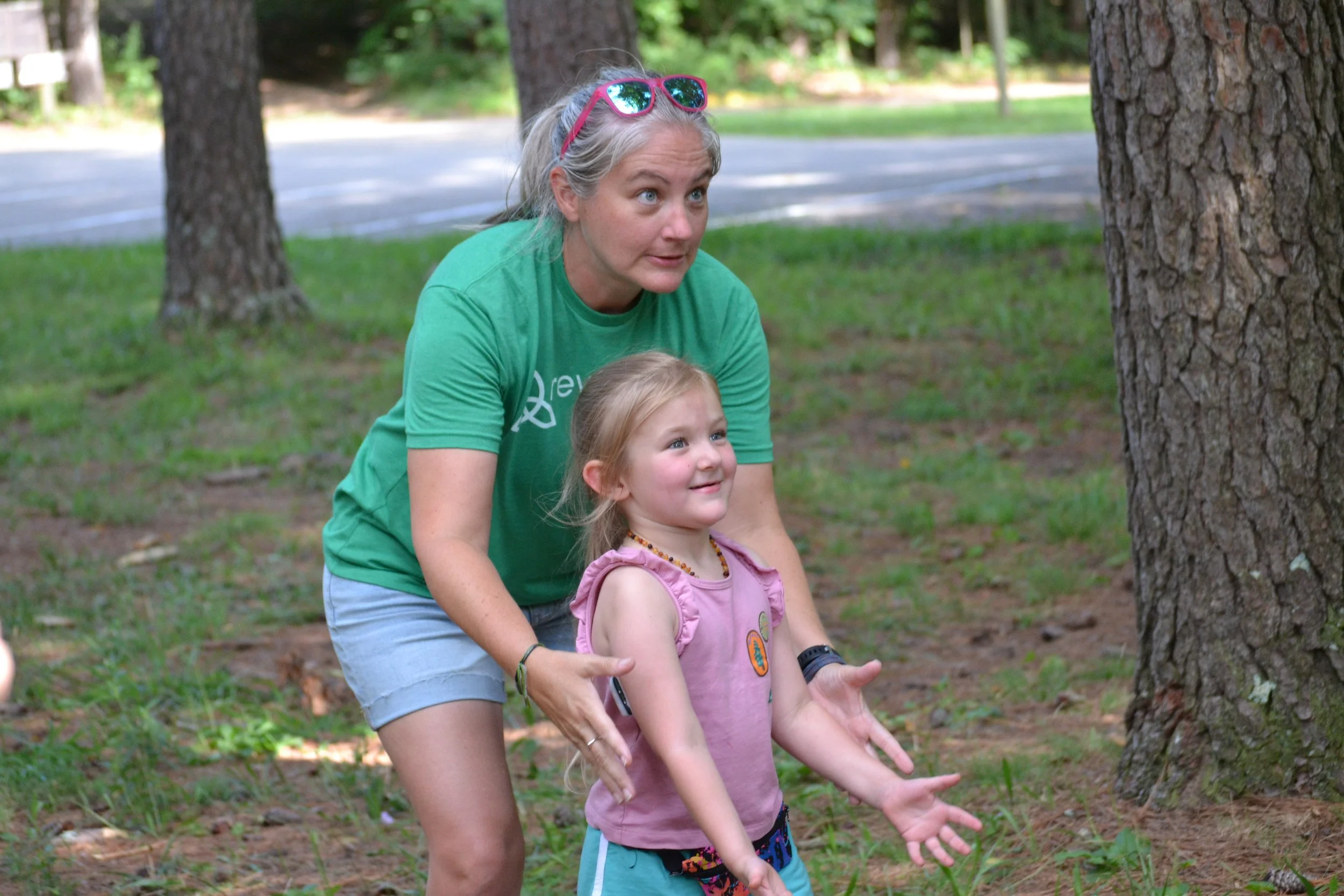 An older woman with sunglasses on her head and a green t-shirt is helping a young girl with blonde hair, pink shirt, and turquoise shorts, stand in a wooded outdoor area with trees and grass.