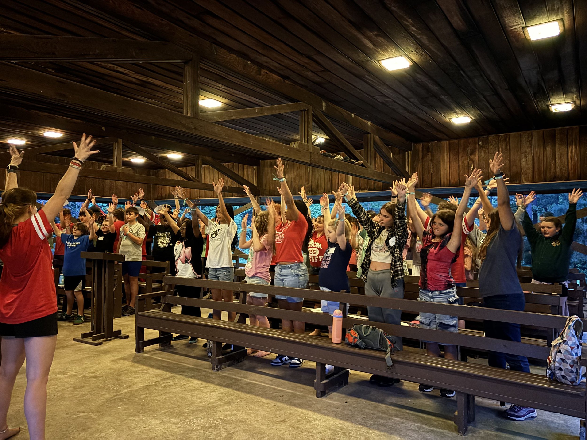 Group of children participating in a standing exercise or activity with their arms raised inside a wooden-paneled room with benches and backpacks.