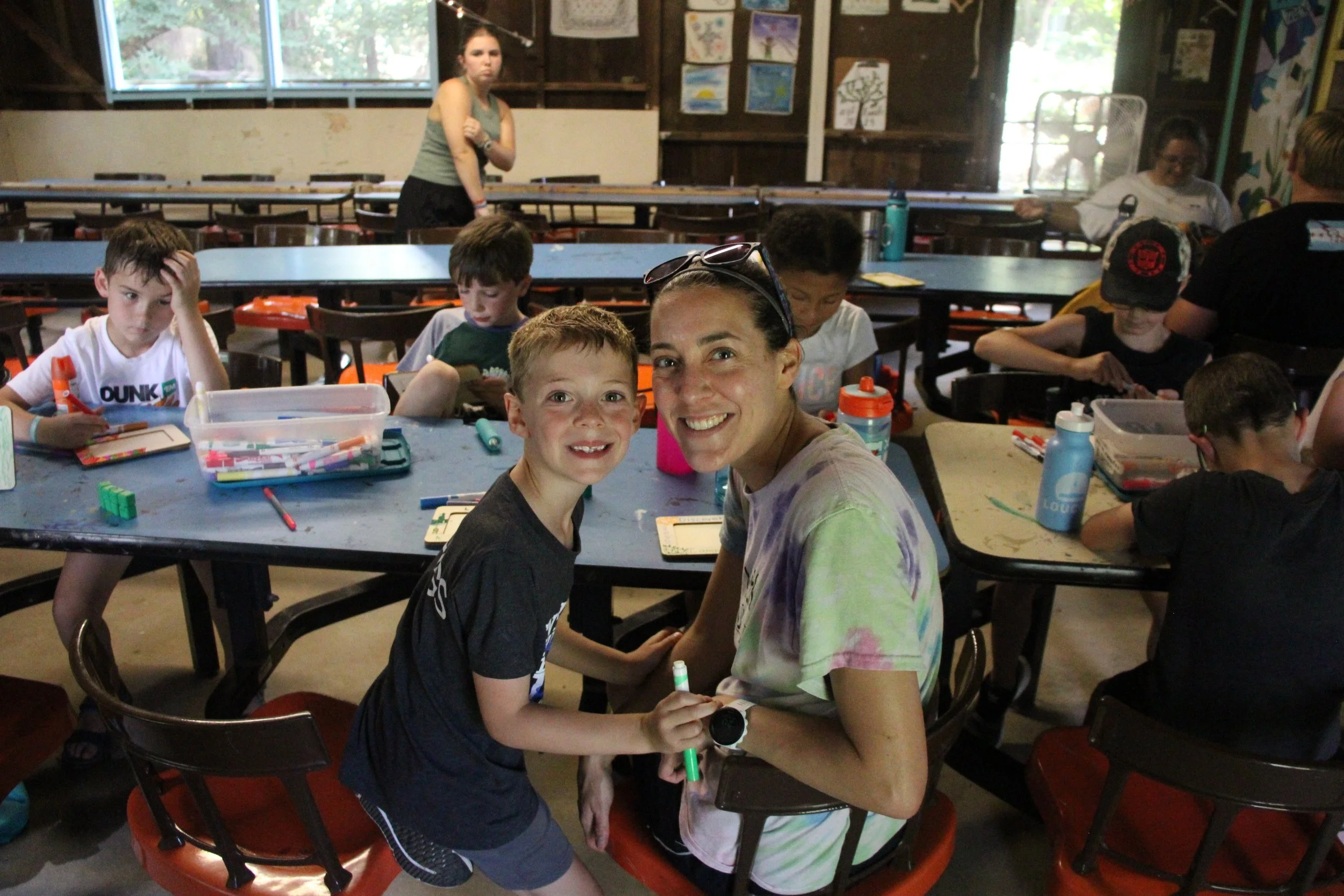 A woman and a young boy smile and hold hands at a table filled with art supplies in a colorful indoor art space with children working on art projects around them.