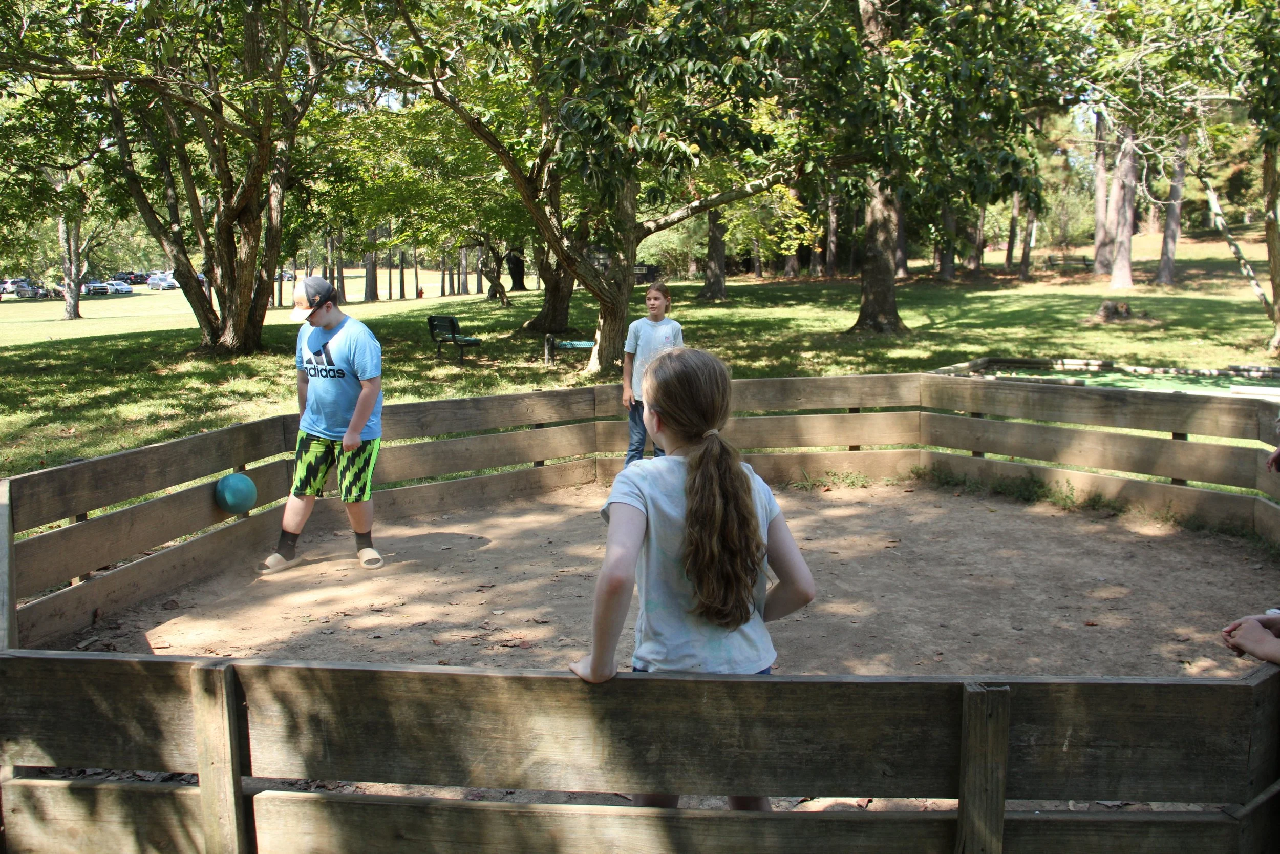 Children playing dodgeball in a fenced dirt circle in a park with trees and grass.