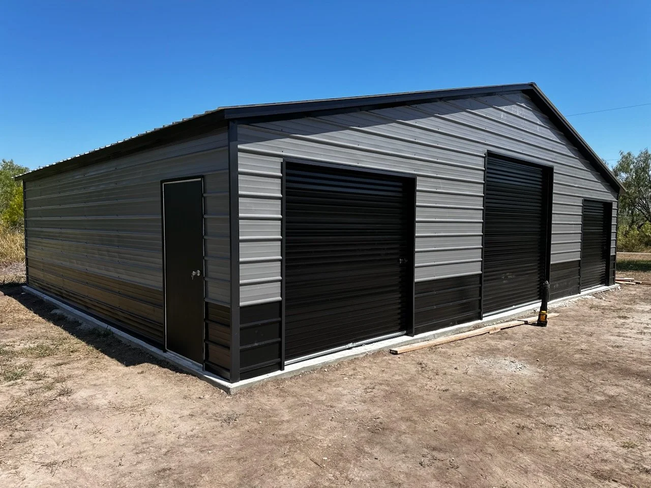 Metal storage building with a sloped roof, single door, and three black roll-up garage doors on a dirt lot under a clear blue sky.