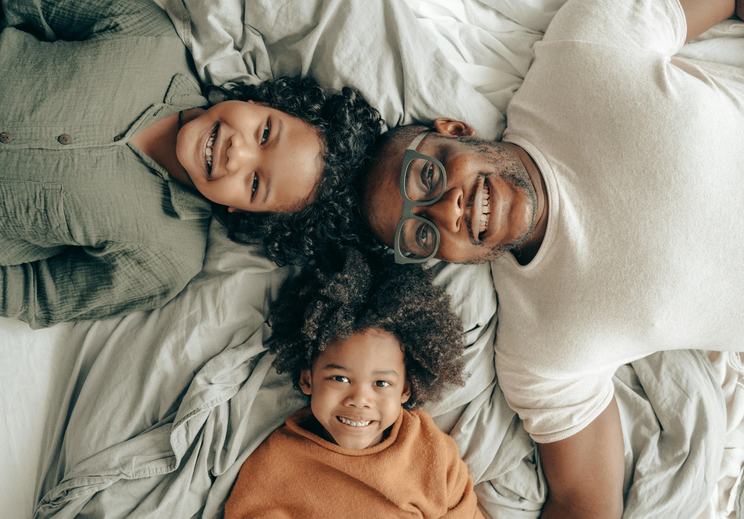 A smiling man with glasses lying on a bed with two young girls, all looking up at the camera.