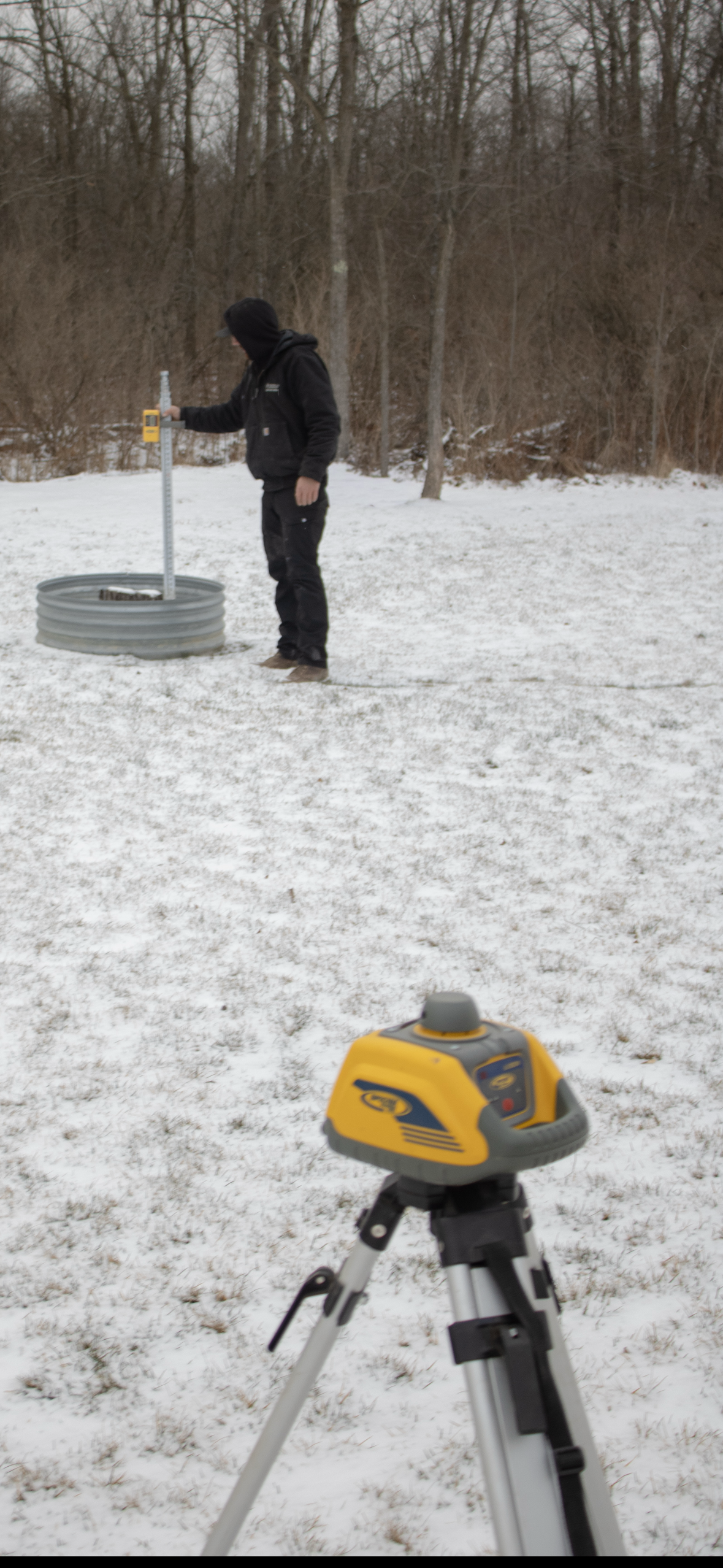 A person using surveying equipment outdoors in a snow-covered field, with a theodolite on a tripod in the foreground and trees in the background.