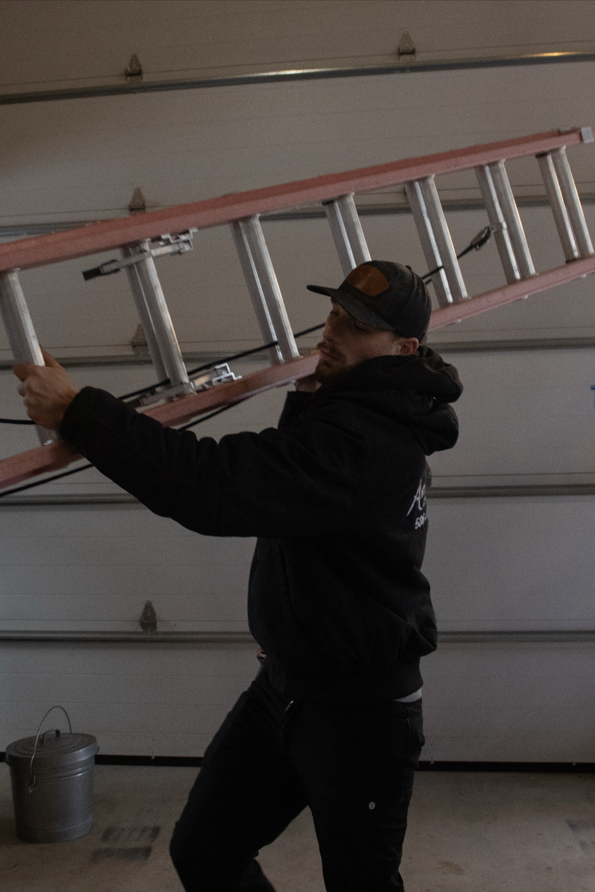 A man wearing a black jacket and a baseball cap is carrying a pink ladder inside a garage.