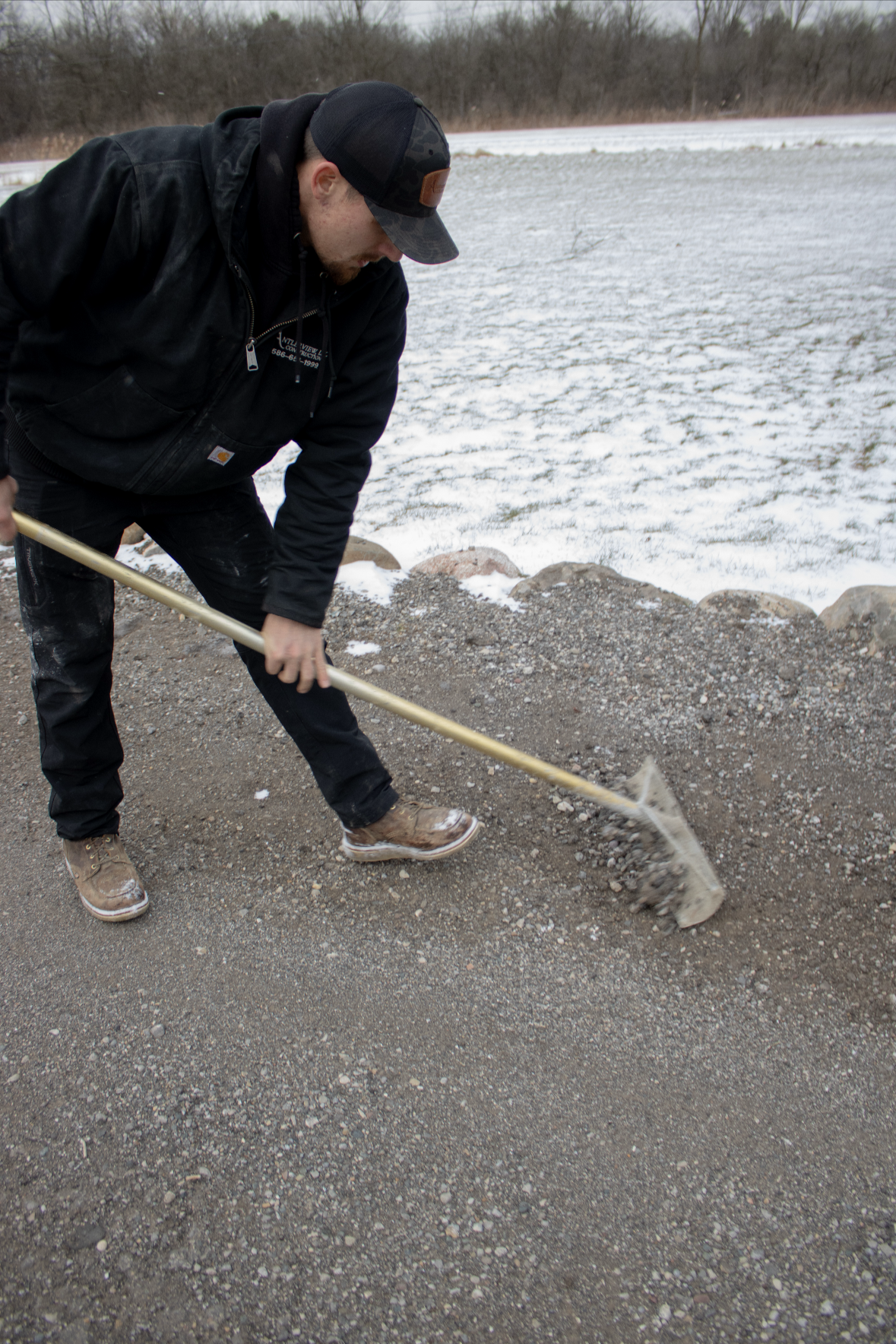 A man wearing a black jacket, black pants, and brown boots is using a rake on a gravel path. The background shows a snow-covered field with bare trees in the distance.