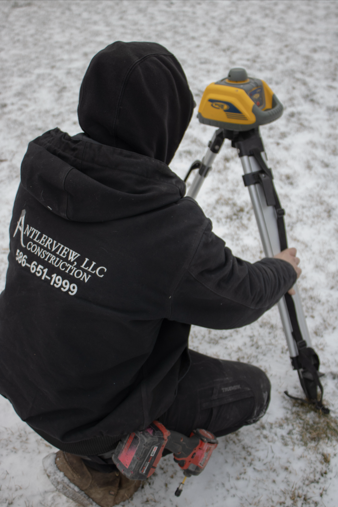 A construction worker kneeling on snow, operating a surveyor device on a tripod.