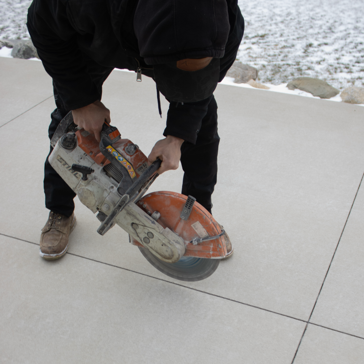 A person using a concrete saw to cut a concrete sidewalk outdoors, with snow and rocks in the background.