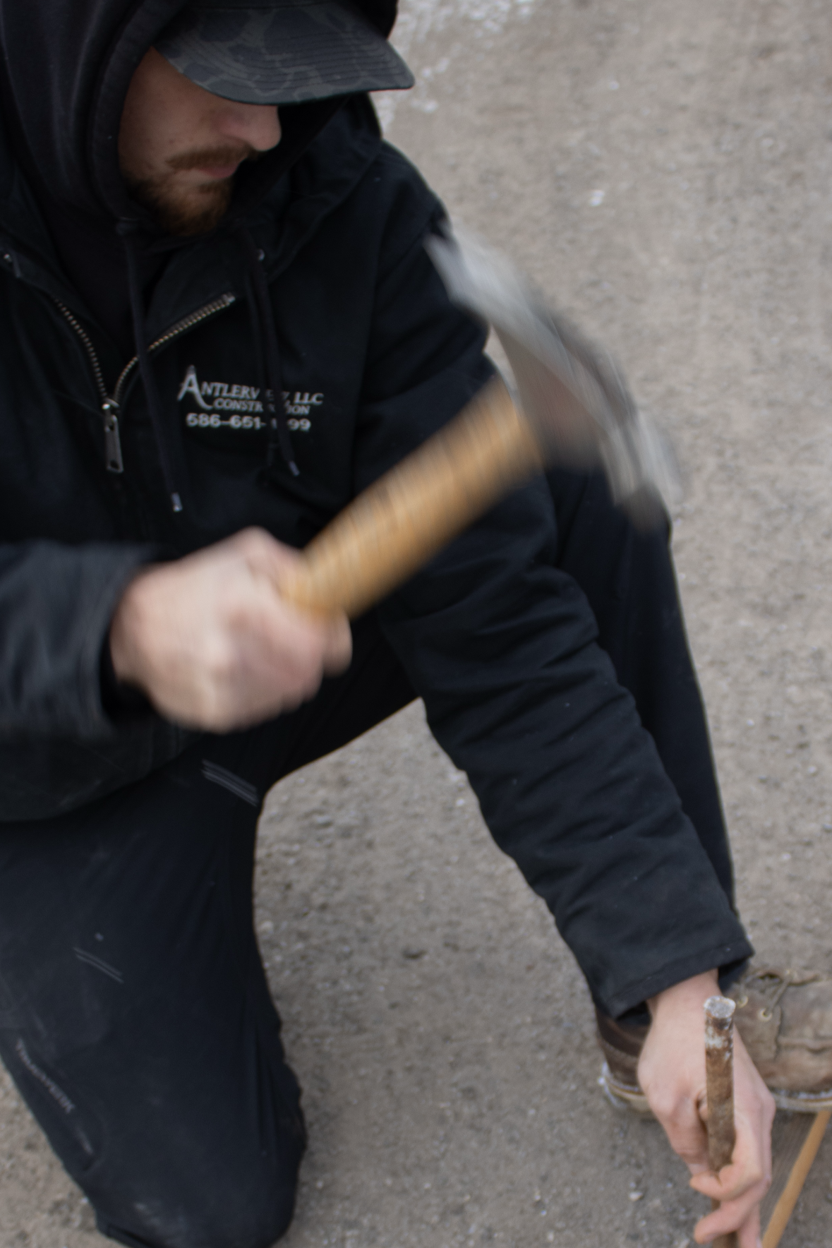 A person wearing a black jacket and cap, crouching down on a dirt surface, holding a hammer in one hand and a metal rod in the other.