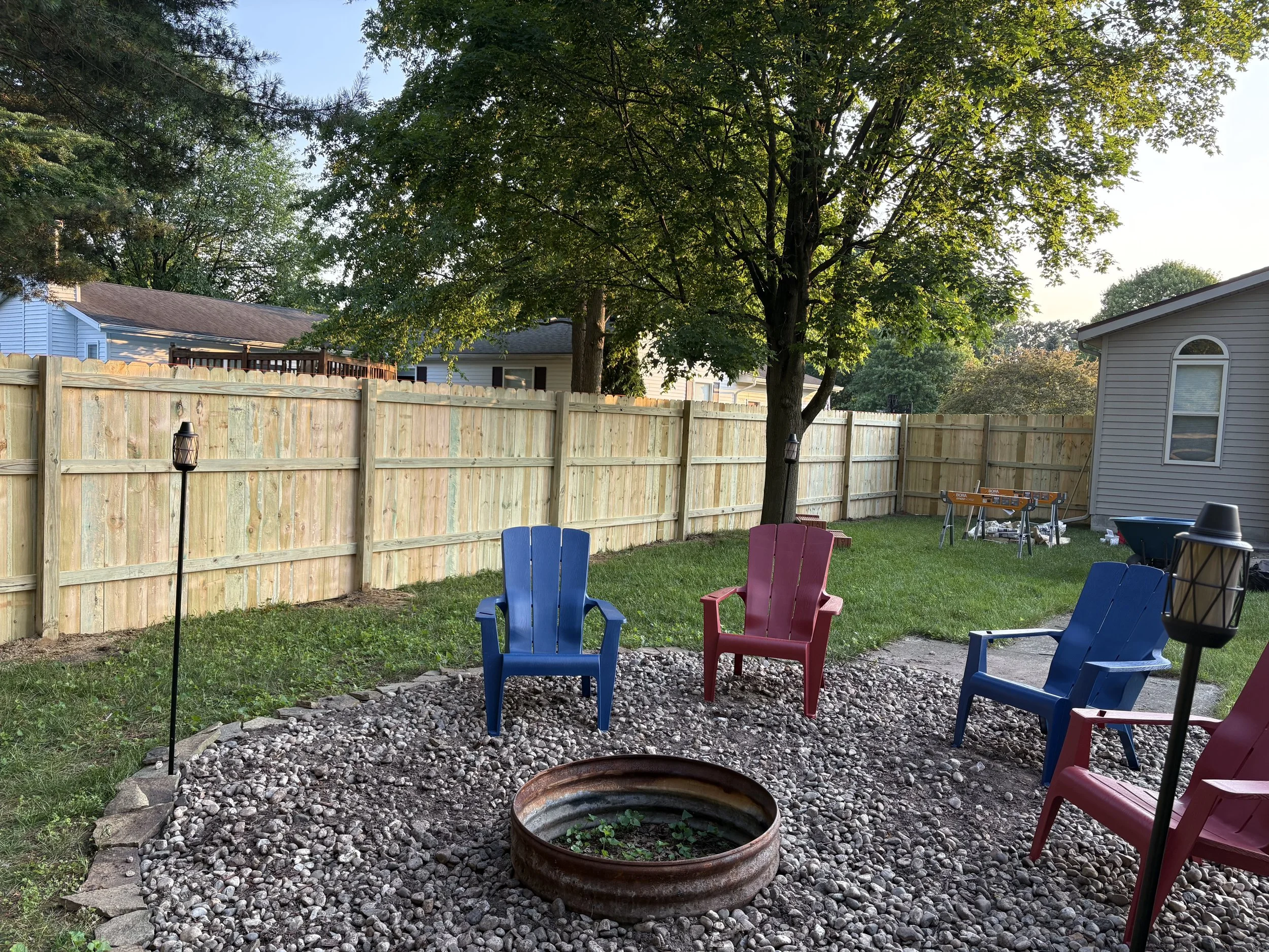 Backyard with colorful Adirondack chairs around a fire pit, wooden fence, trees, and house in late afternoon sunlight.