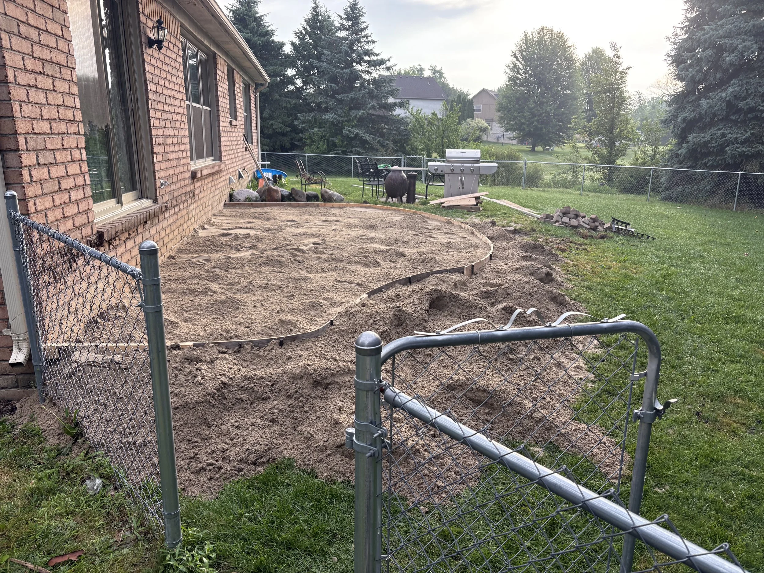 Backyard with a sand or soil area being prepared for a patio or garden. A partial fence surrounds the area, and there are trees, a barbecue grill, and outdoor furniture in the background.