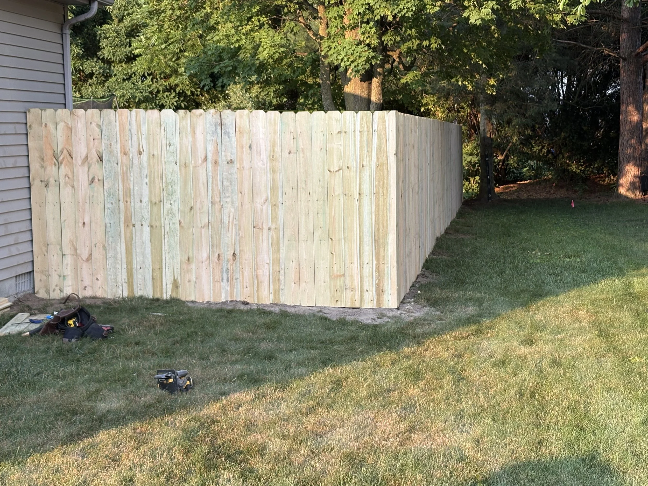 A freshly built wooden privacy fence adjacent to a house, with tools and supplies on the grass nearby, and a background of trees and a grassy yard.