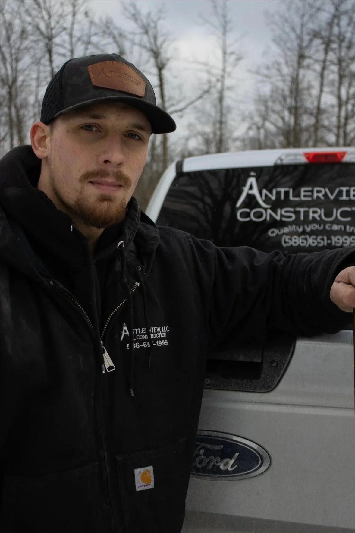 A man with a beard and mustache wearing a black hoodie and a trucker hat, standing next to a light-colored Ford truck with a company logo, in a wooded area with bare trees in the background.