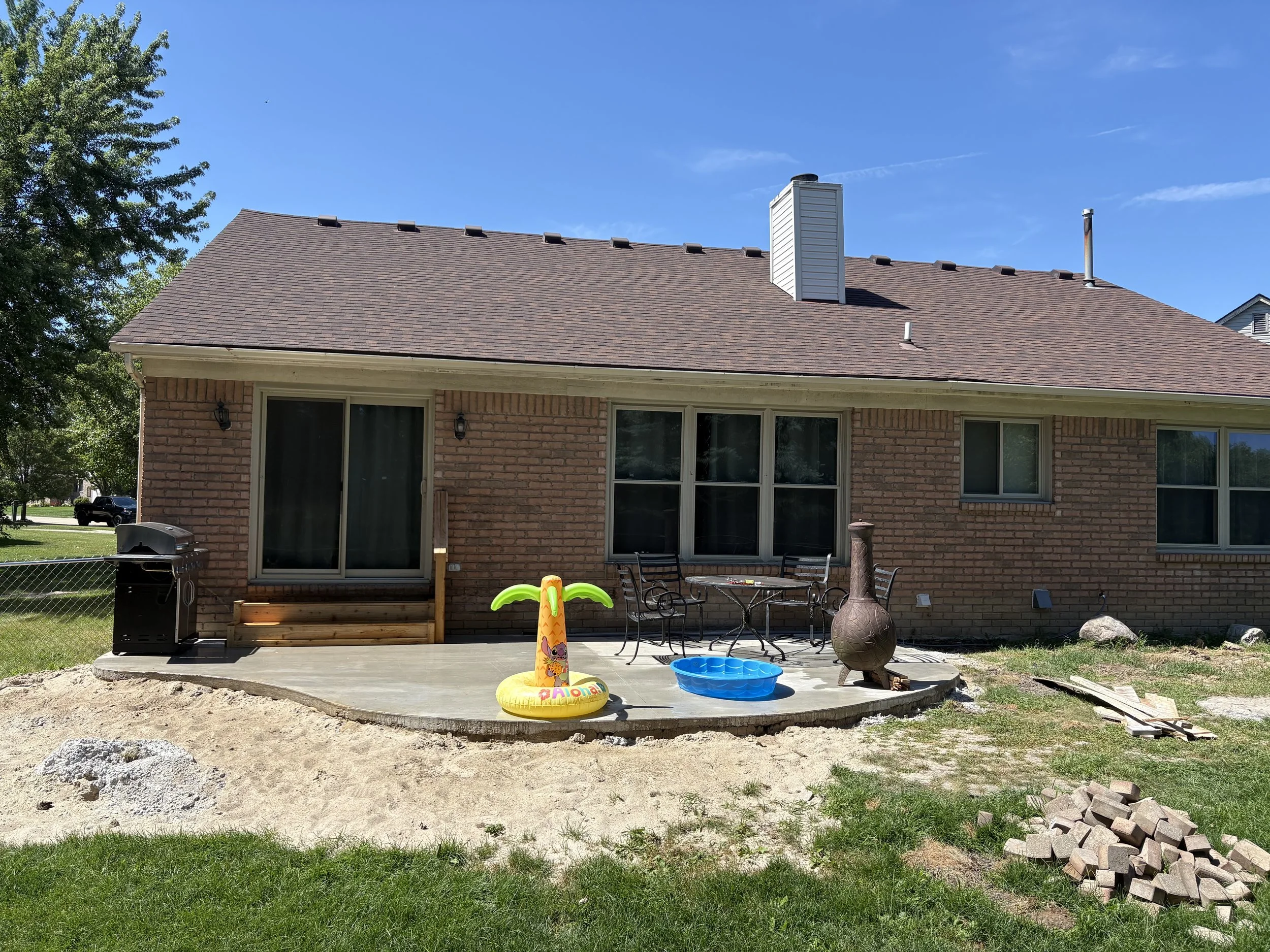 Backyard with a patio, children's inflatable pool, outdoor furniture, a grill, and a chimney, set against a red brick house with sliding door and windows under a blue sky.