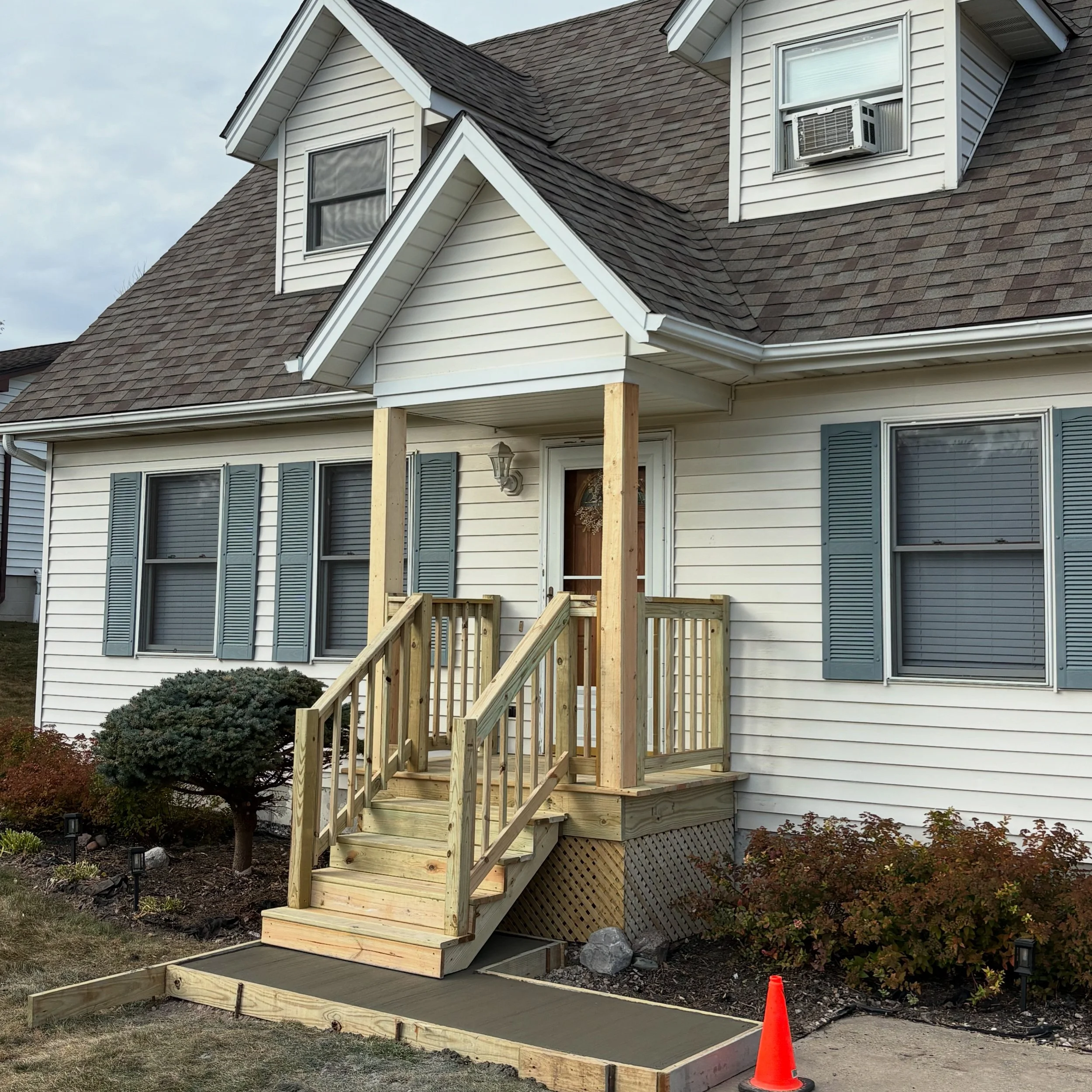 Front porch new wooden stairs and railing under construction on white house with blue shutters, surrounded by bushes and a small landscaped yard, orange construction cone on sidewalk.