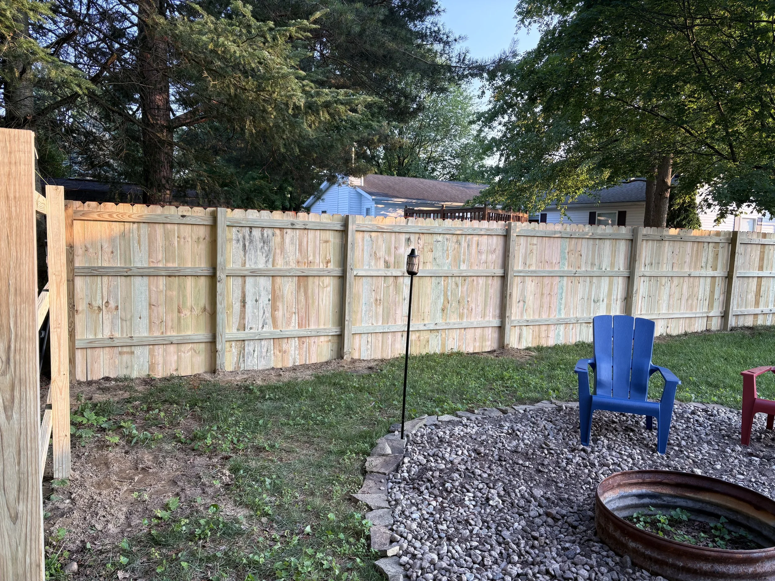 A backyard with a wooden privacy fence, two colorful Adirondack chairs (blue and red), a small garden bed with rocks and an old metal wheel, a lamp post, tall trees, and neighboring houses in the background.