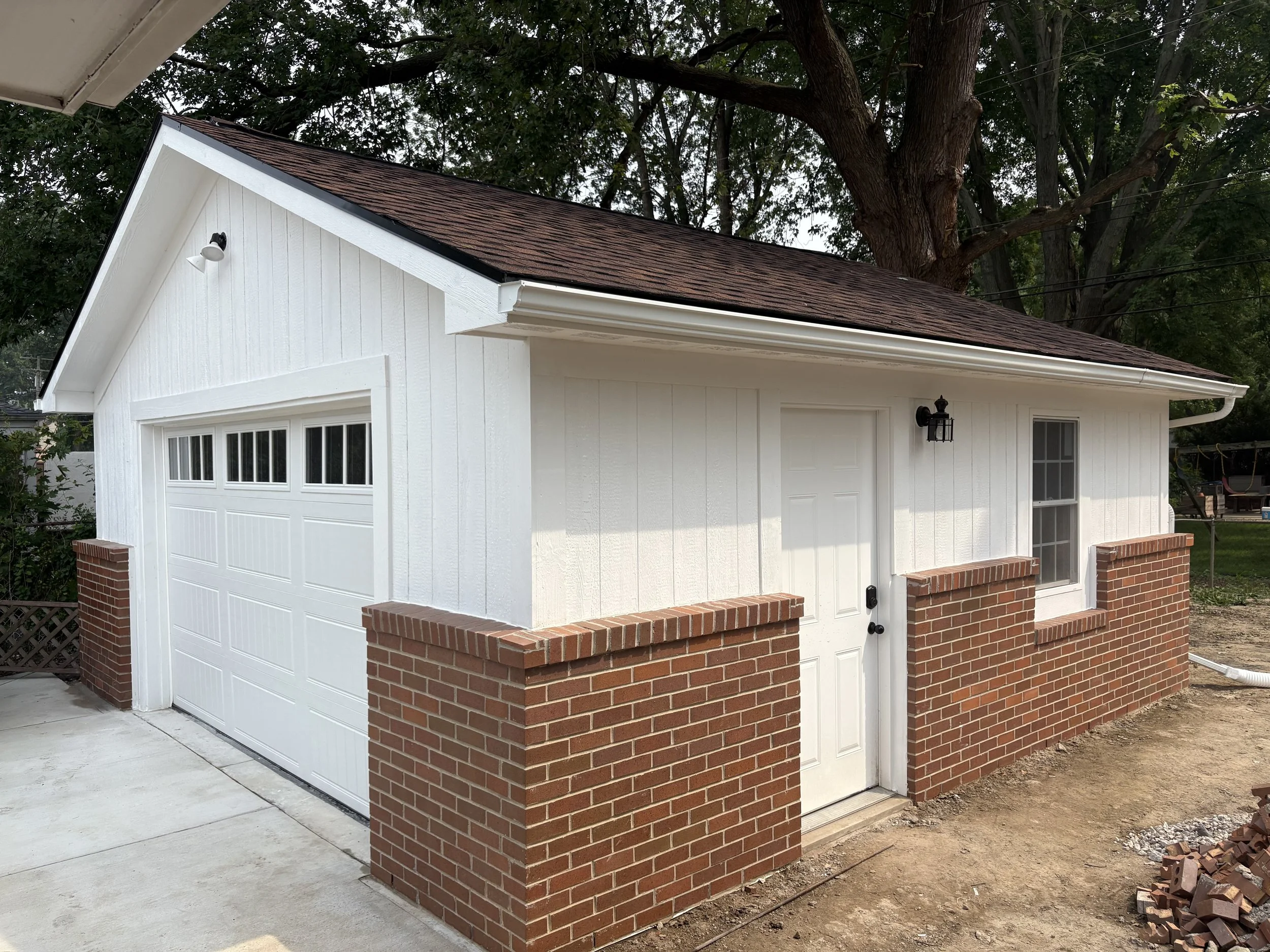 A small white garage with a brick walled base, a white door, and a new brick wall on the front. There are two windows with grid patterns, and a black outdoor light fixture. The roof is brownish with shingles, and there are trees in the background.