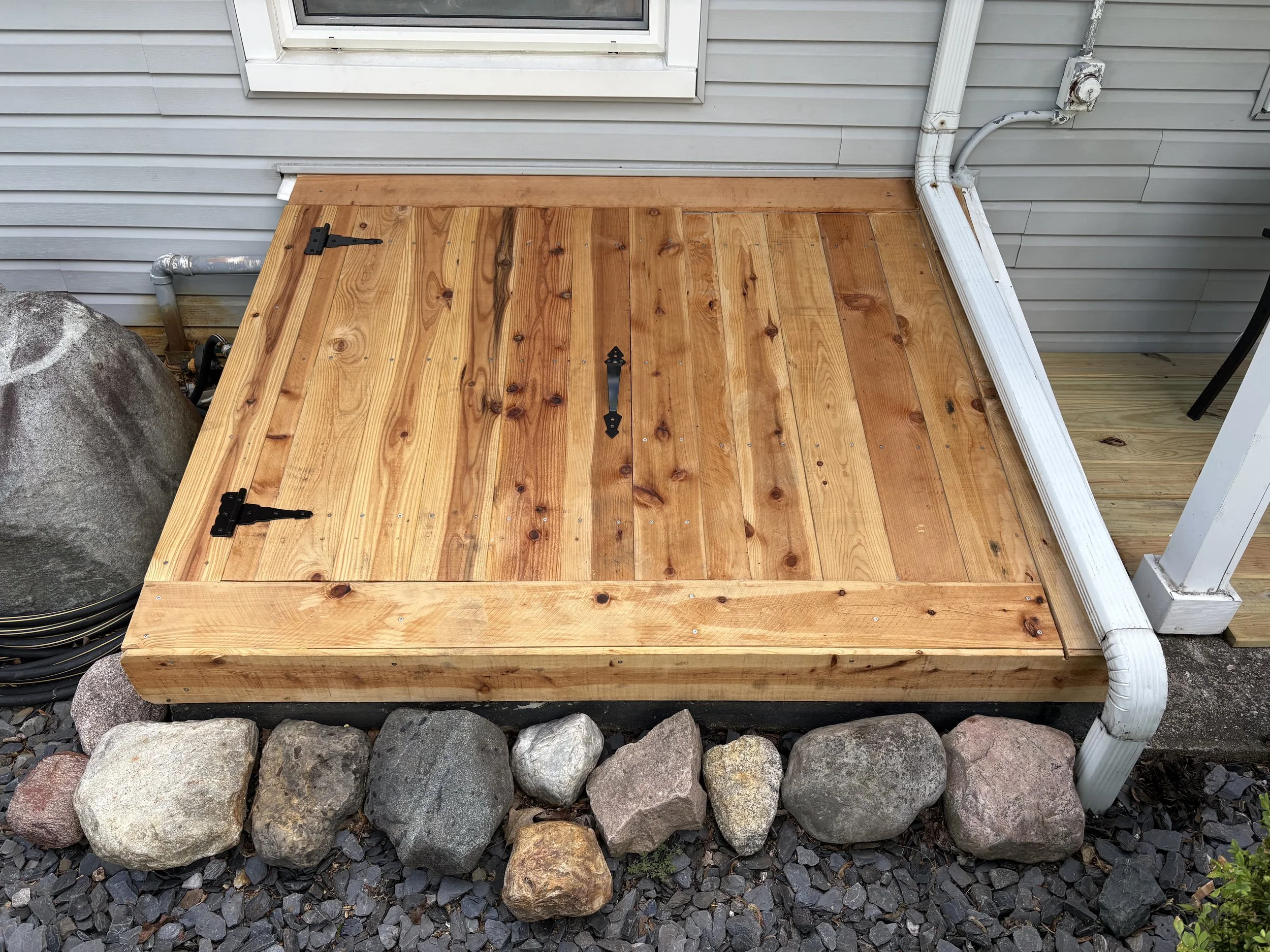 Freshly built wooden deck with black metal hinges and a handle, surrounded by rocks and gray gravel, attached to a house with gray siding and white trim.