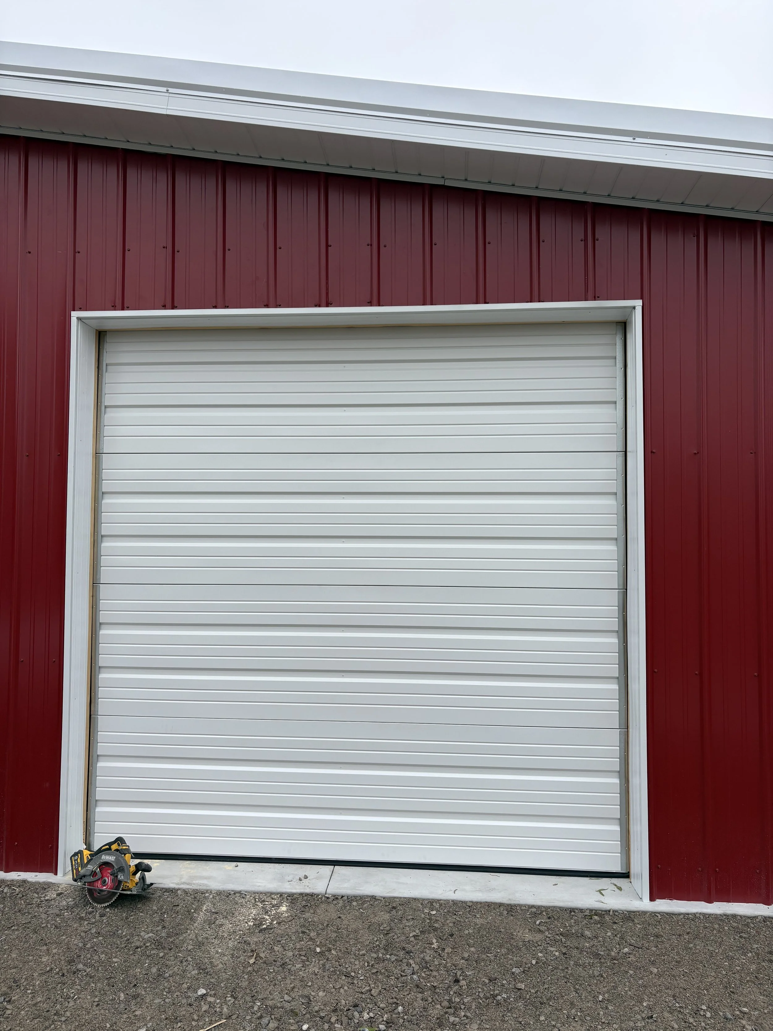 A white garage door on a red building with a circular saw placed on the ground to the left side.