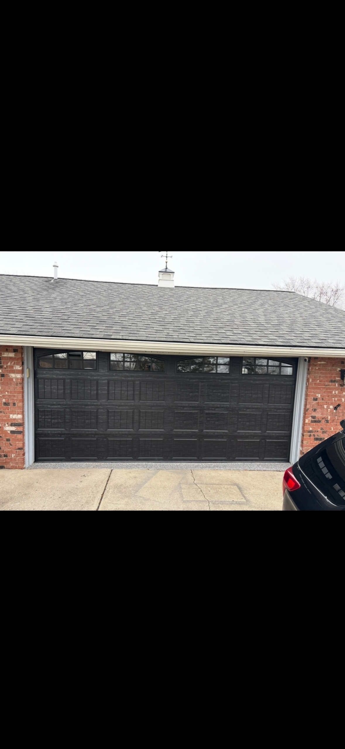 Black garage door with small windows at the top, brick walls on either side, gray roof, and part of a black car visible on the lower right.