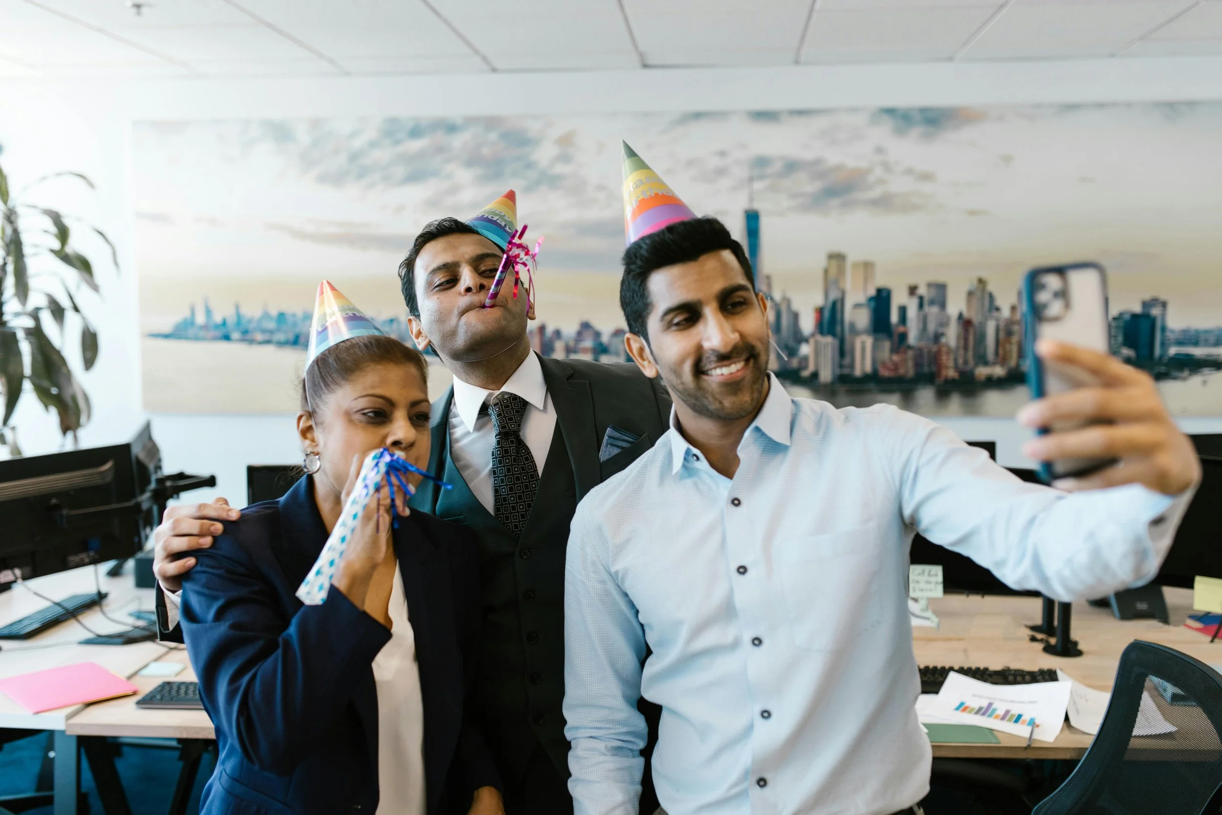 Three colleagues celebrating a birthday in an office, wearing party hats, with confetti and a selfie being taken.