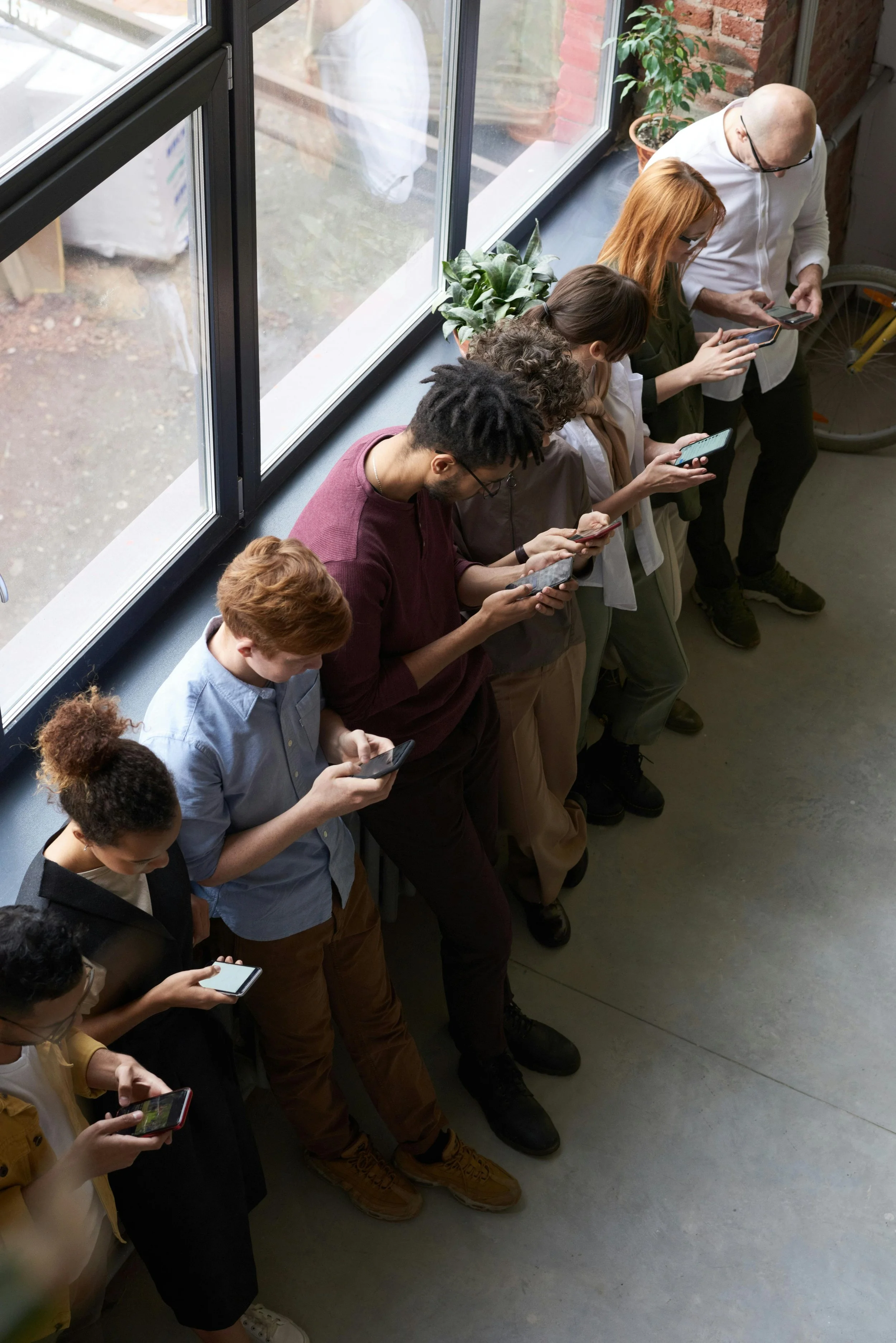 A group of people standing in a line, all looking at their smartphones, by a large window in an indoor setting.