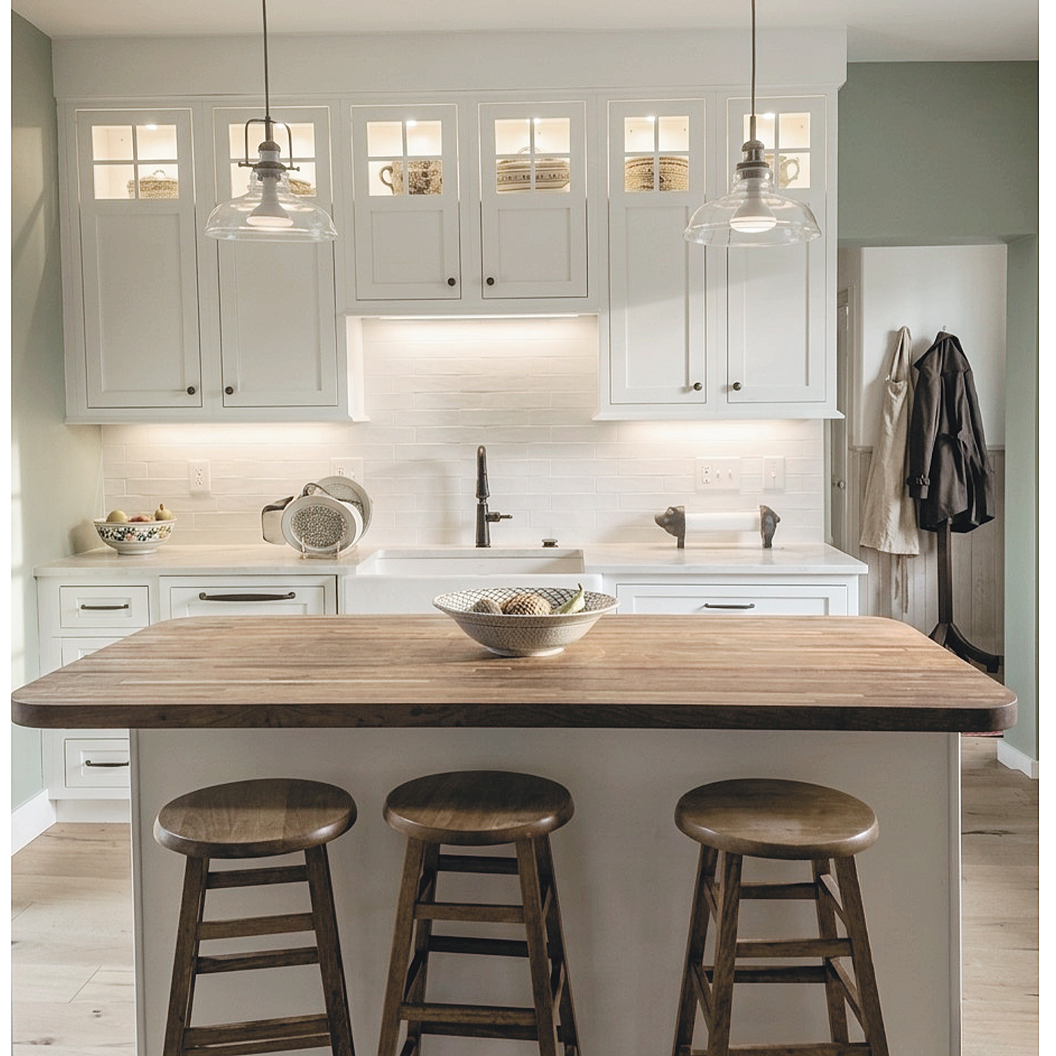 A modern kitchen with white cabinetry, a farmhouse sink, pendant lights, and a butcher block island with three wooden stools. A bowl with decorative balls is on the island, and a coat rack with hanging coats is in the background.