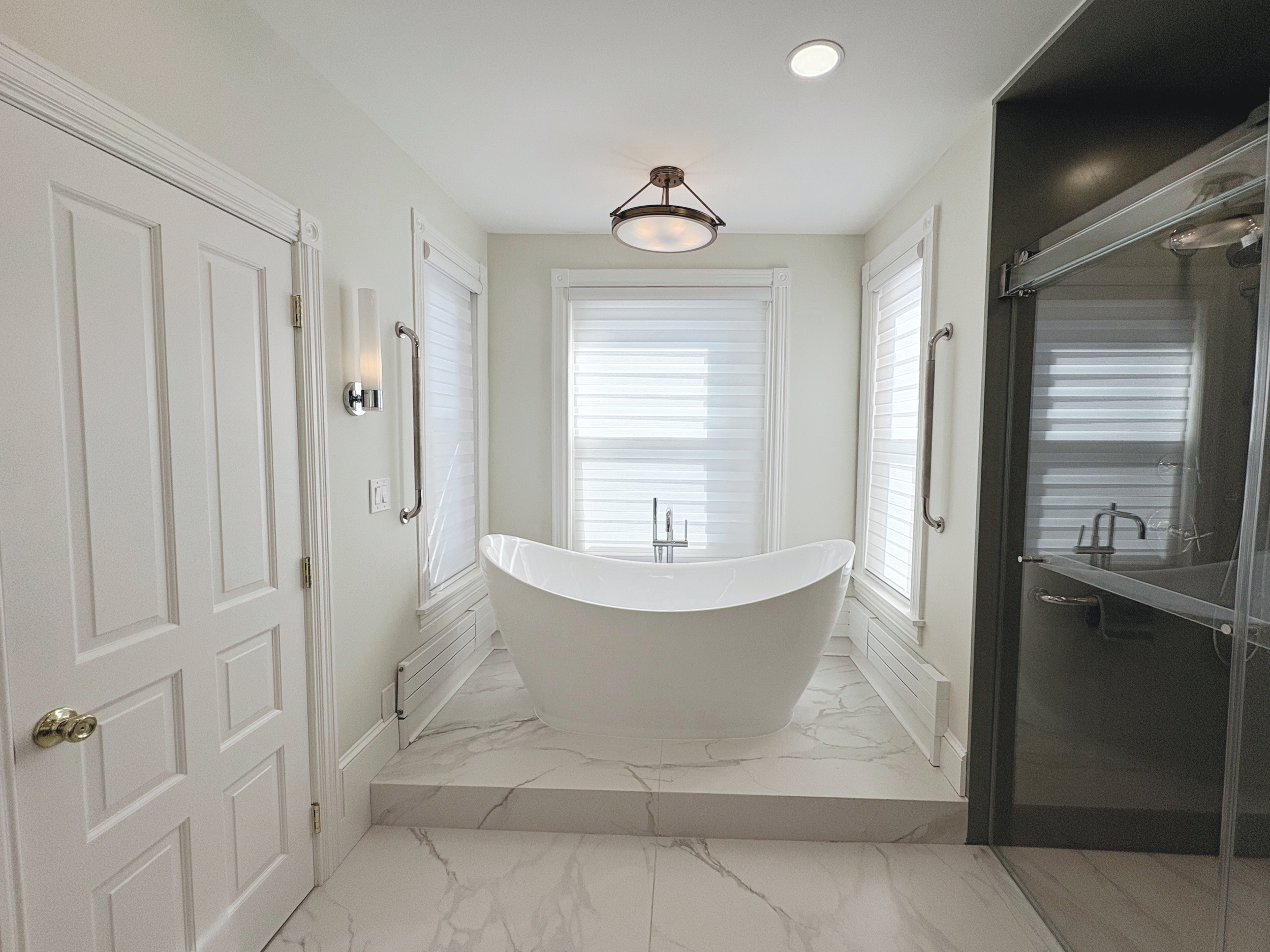 A bathroom with a white freestanding bathtub in front of three large windows with white blinds, a black shelving unit on the right, and white doors on the left. The floor and platform are tiled with white marble. The ceiling has a central round light