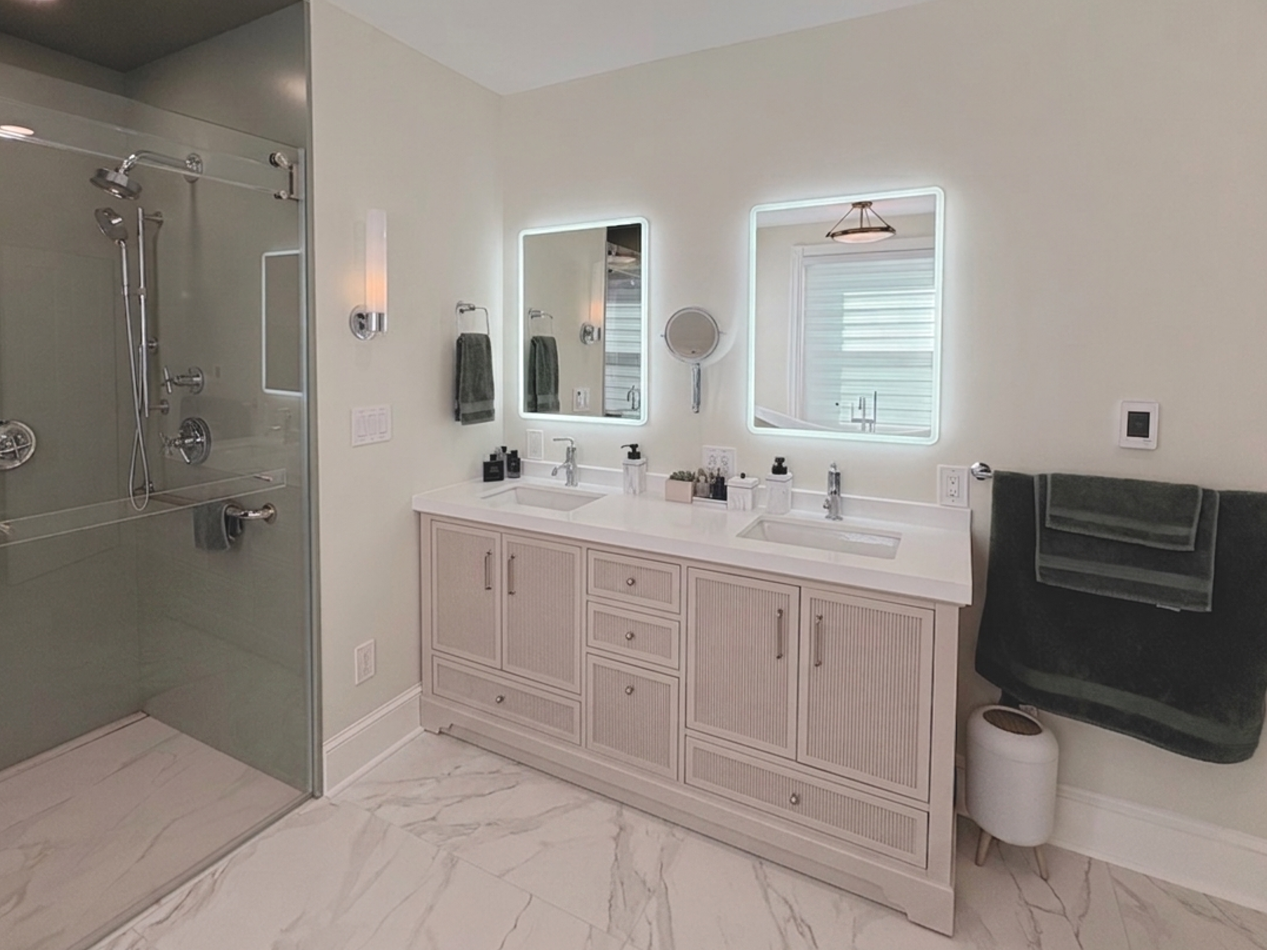 Modern bathroom with walk-in shower, double vanity sinks, illuminated mirrors, dark gray towel, and marble floor.