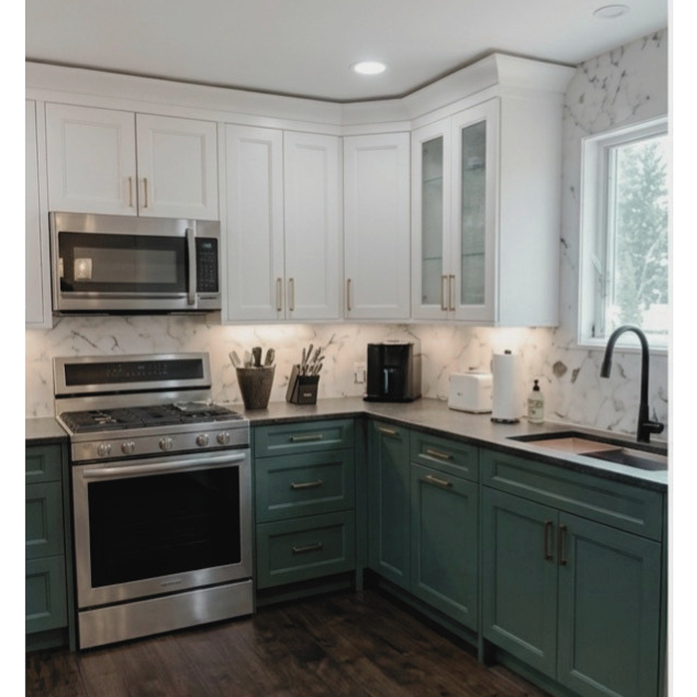 Modern kitchen with white upper cabinets, green lower cabinets, stainless steel stove and microwave, black faucet, marble backsplash, and a window above the sink.