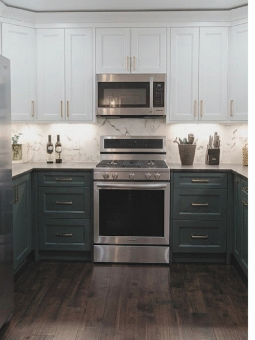 Modern kitchen with white upper cabinets, navy lower cabinets, stainless steel appliances including a microwave installed above a stove, marble backsplash, and dark wooden flooring.