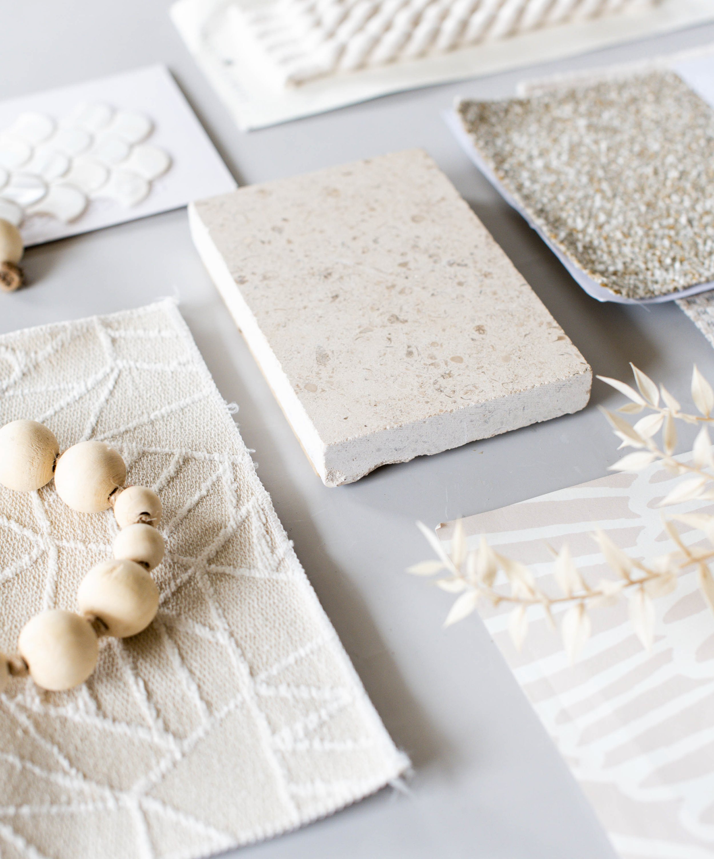 Close-up of texture and color samples including a beige stone tile, speckled tile, and textured fabric with a string of white beads and dried leaves nearby.