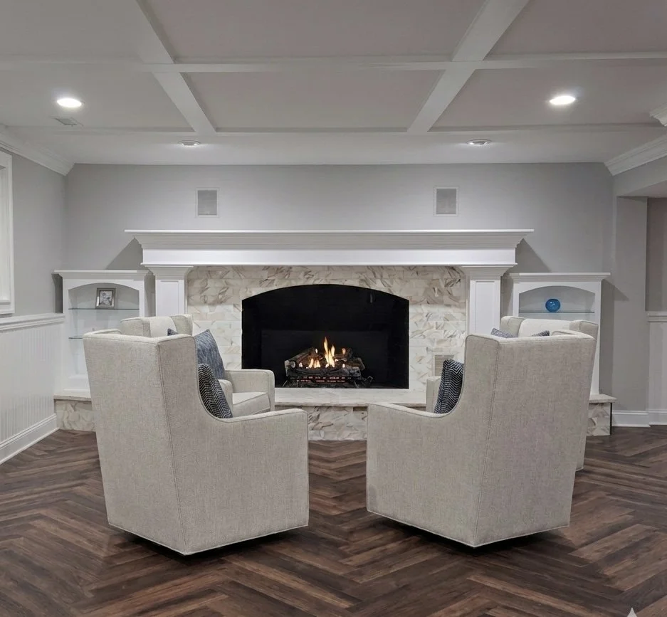 Cozy living room with two beige armchairs facing a fireplace, built-in shelves on each side, and a white mantel. The fireplace has a fire burning inside, and the room has a neutral color palette with gray walls and dark wood flooring.