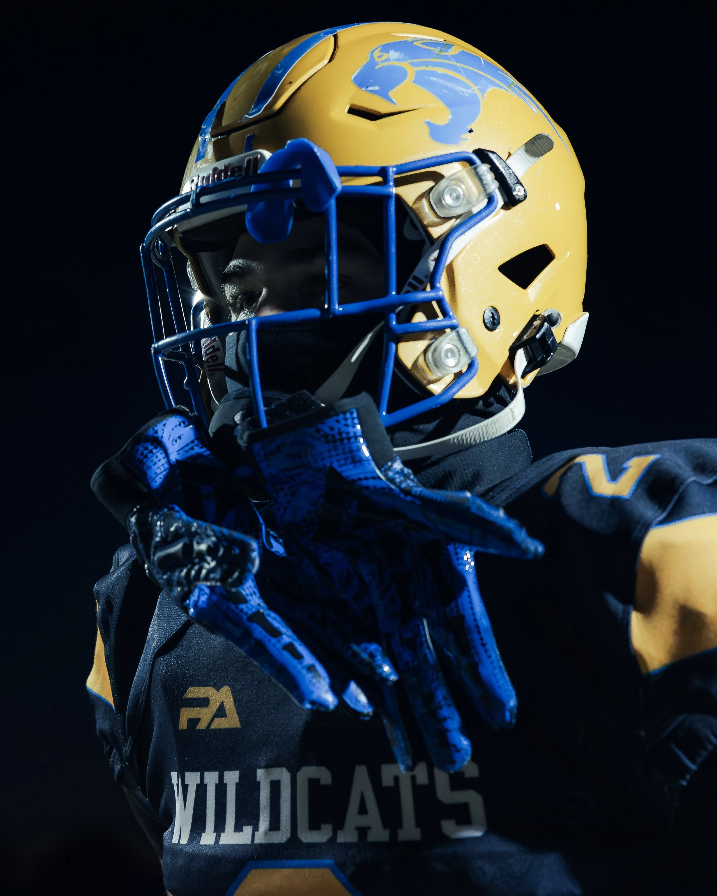 A Wildcats football player in a yellow helmet adjusts a blue glove under dramatic lighting.