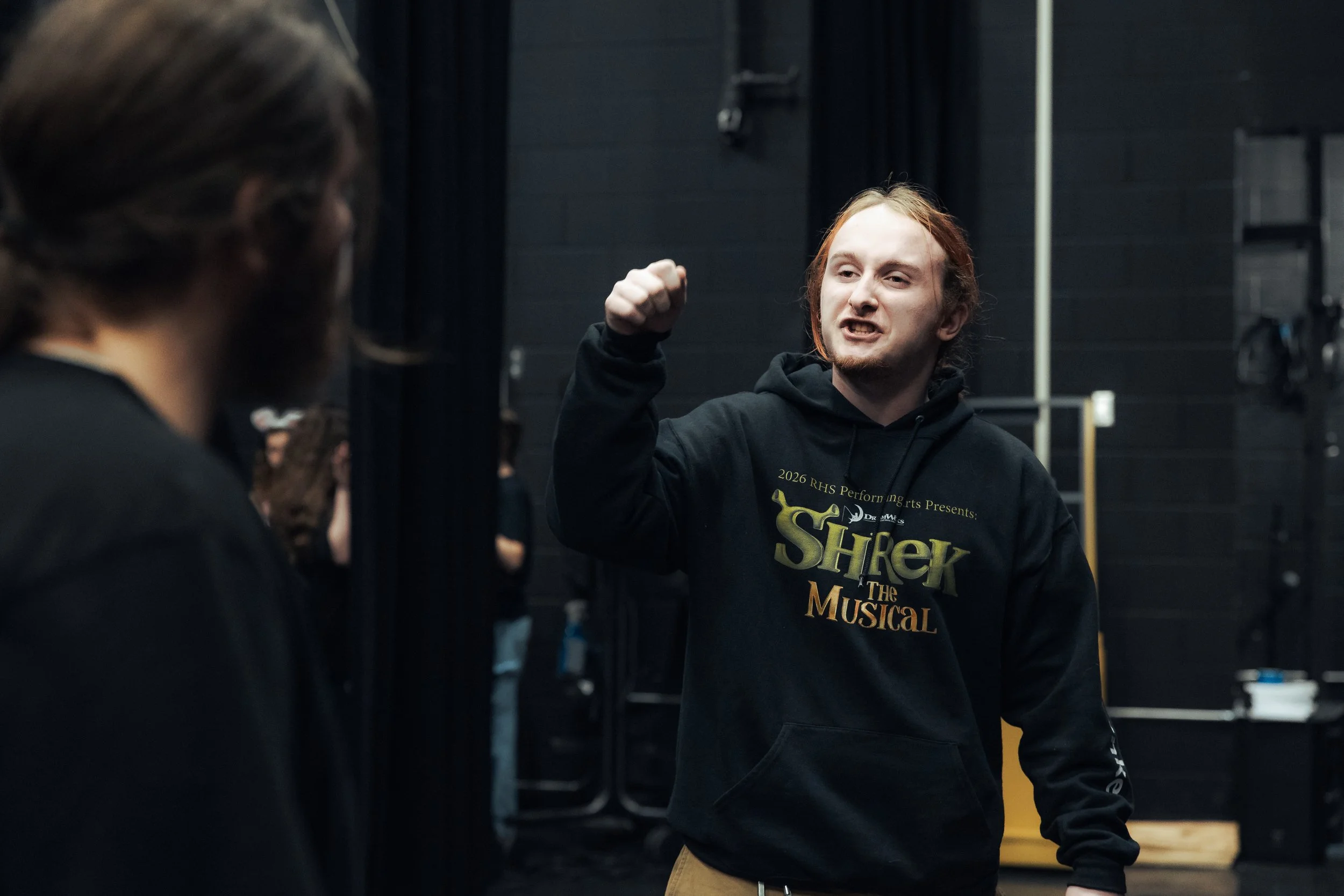 A cast member gestures during rehearsal near the stage curtains.
