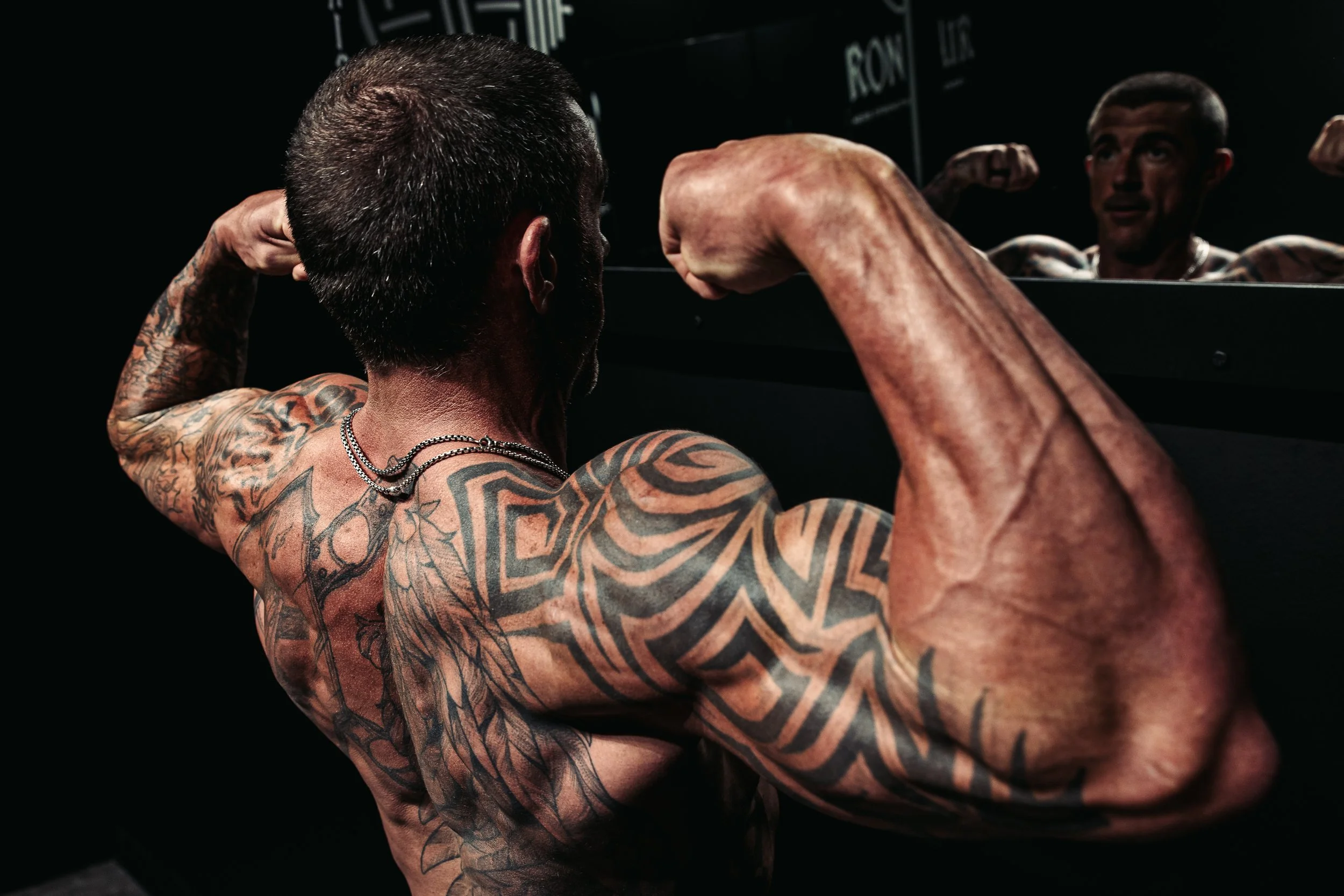 Rear view of a tattooed bodybuilder posing in a gym mirror.