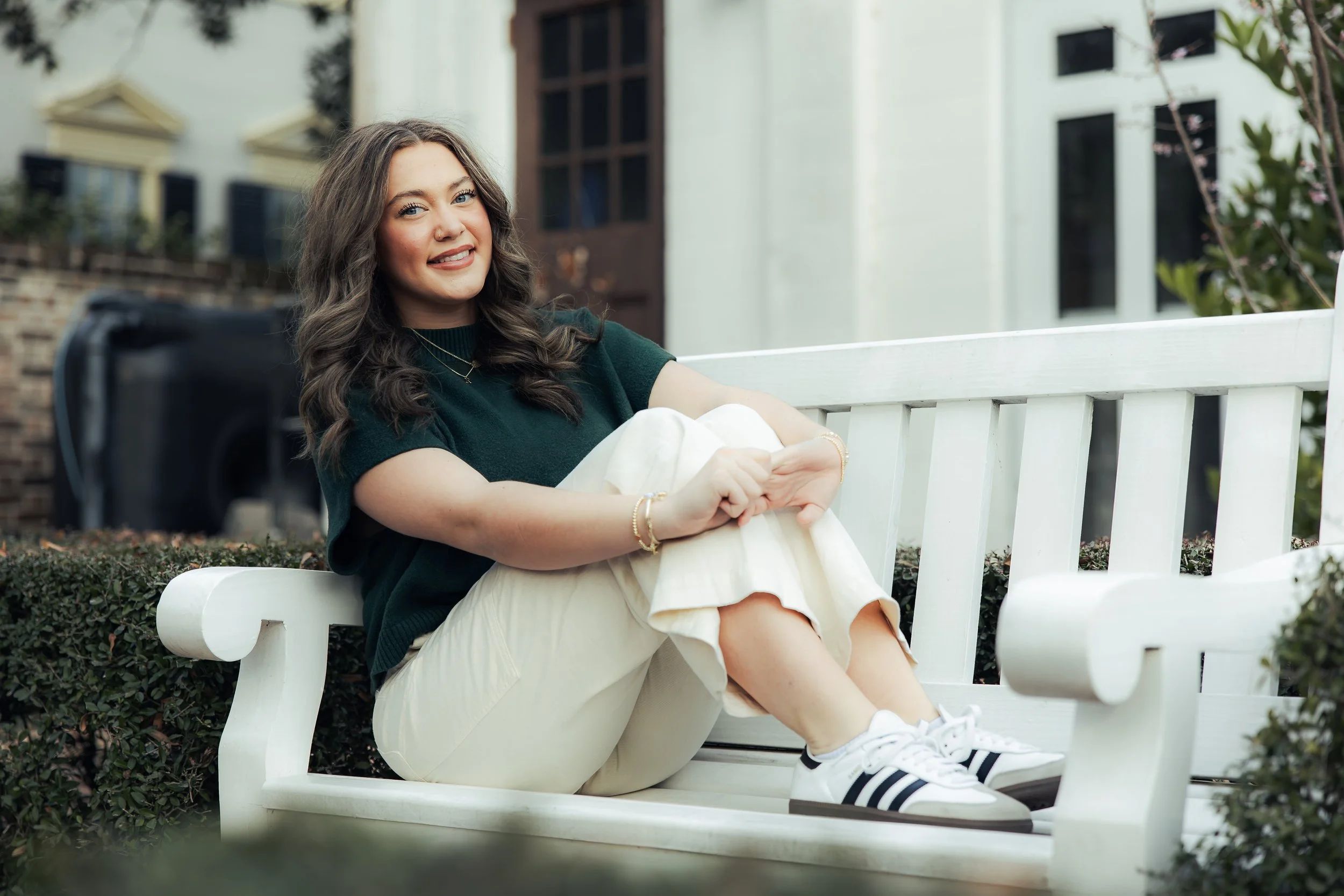 A young woman in a green sweater and cream pants sitting on a garden bench.