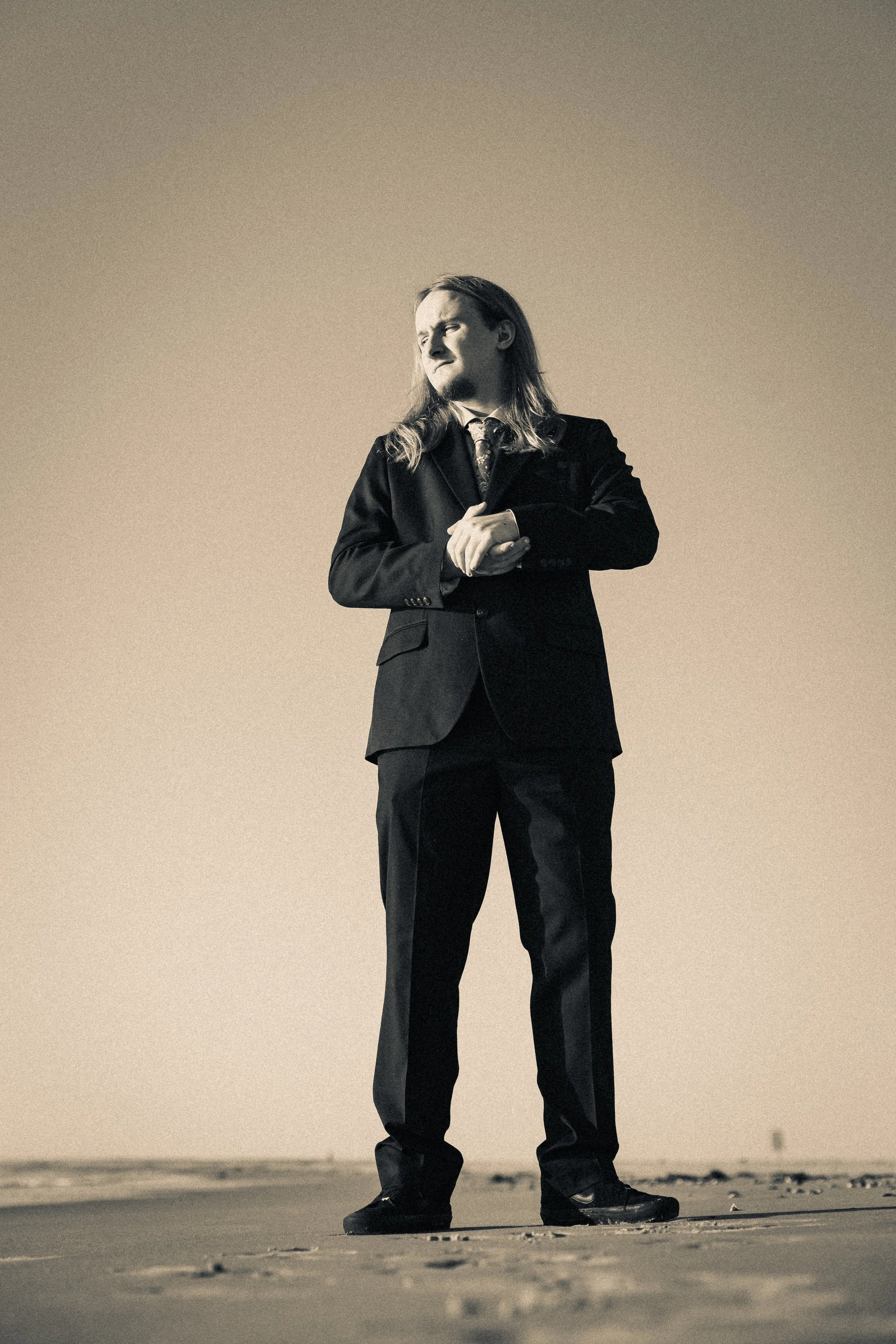A man in a black suit adjusting a cuff on the beach.