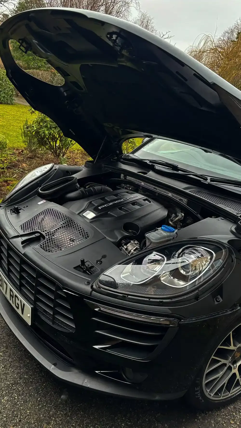 Front of a black Porsche with the hood open, showing the engine, parked on a driveway with grass and trees in the background.