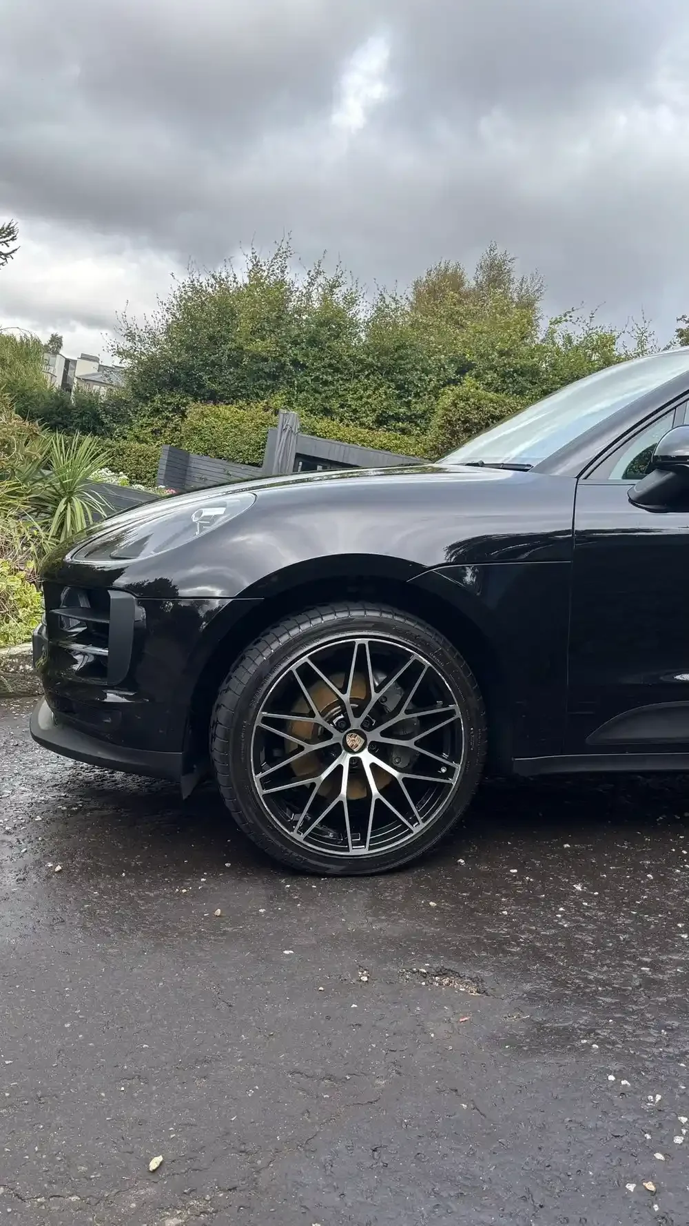 Close-up of front part of a black Porsche car parked on asphalt with greenery and cloudy sky in the background.