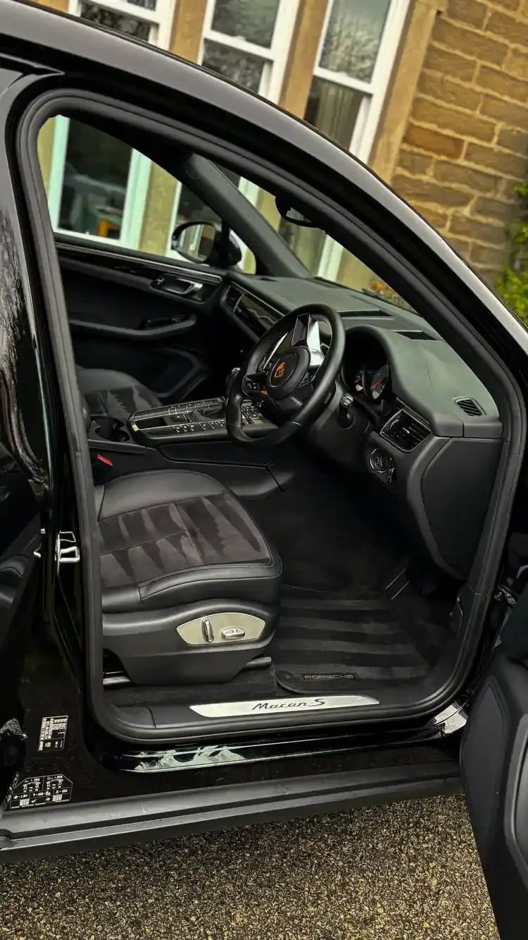 Interior of a black Porsche Macan S with leather seats and a dashboard, viewed from the open driver-side door, outside a house with brick walls and large windows.