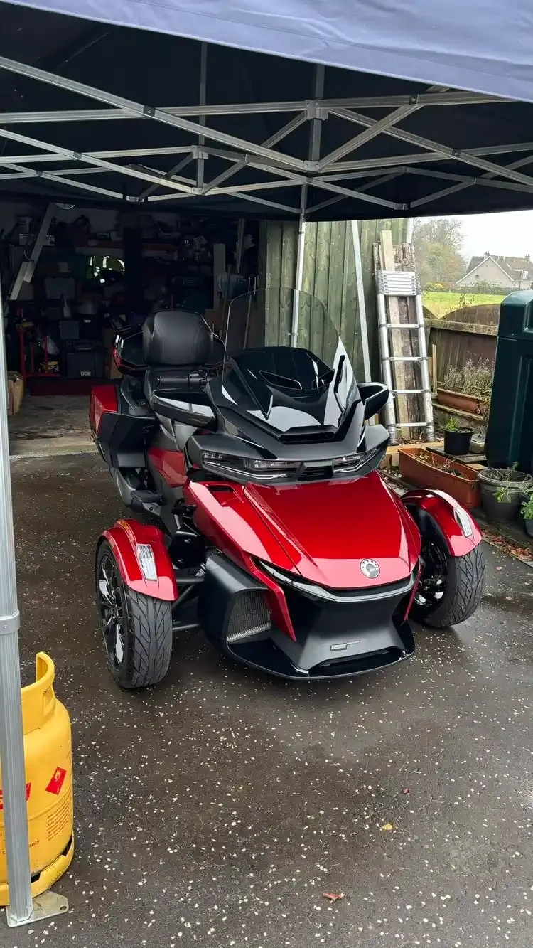 Red and black Can-Am Spyder three-wheeled motorcycle parked under a canopy in a driveway.