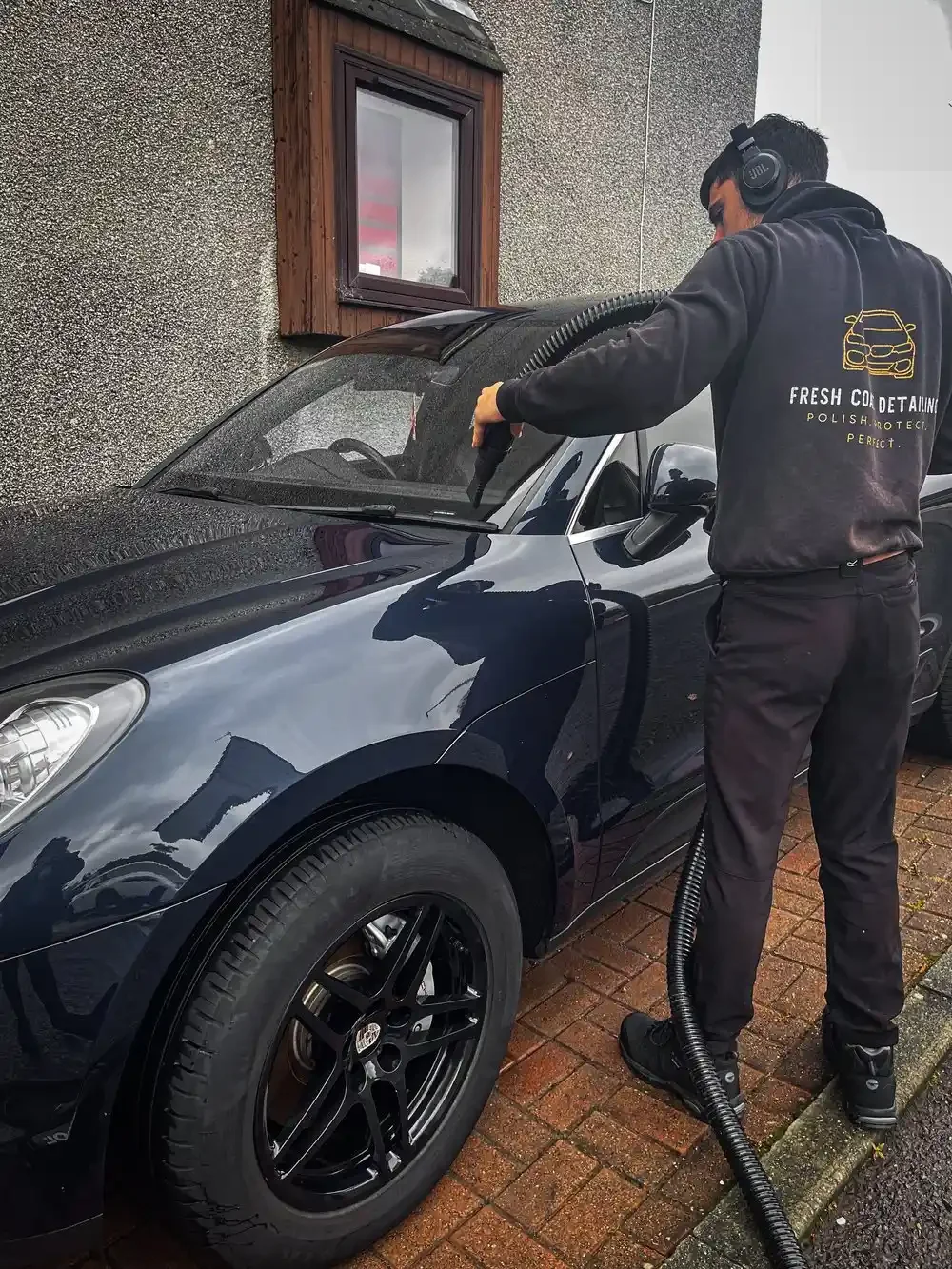Man wearing headphones and branded hoodie using a water hose to wash a black car parked on a brick pavement beside a textured wall and window.
