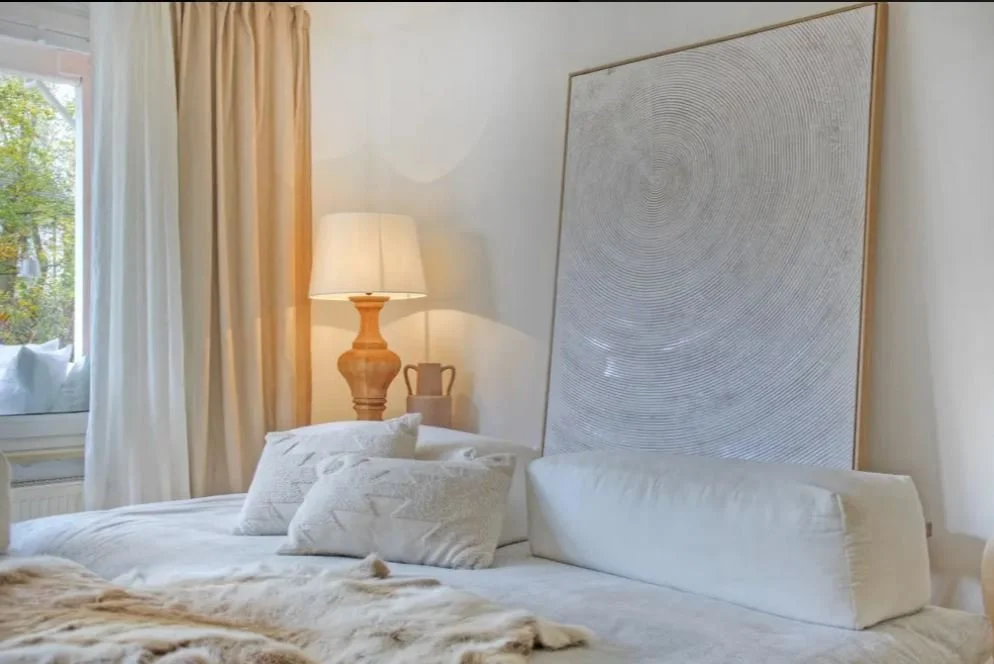 Geräumiges Schlafzimmer mit weißem Bett, Kissen, einem großen Rahmen mit blassgrauem grafischem Muster, einem Holzlampenschirm, einer Tischvase und Blick auf ein geöffnetes Fenster mit gelben Vorhängen, durch das grüne Bäume zu sehen sind.