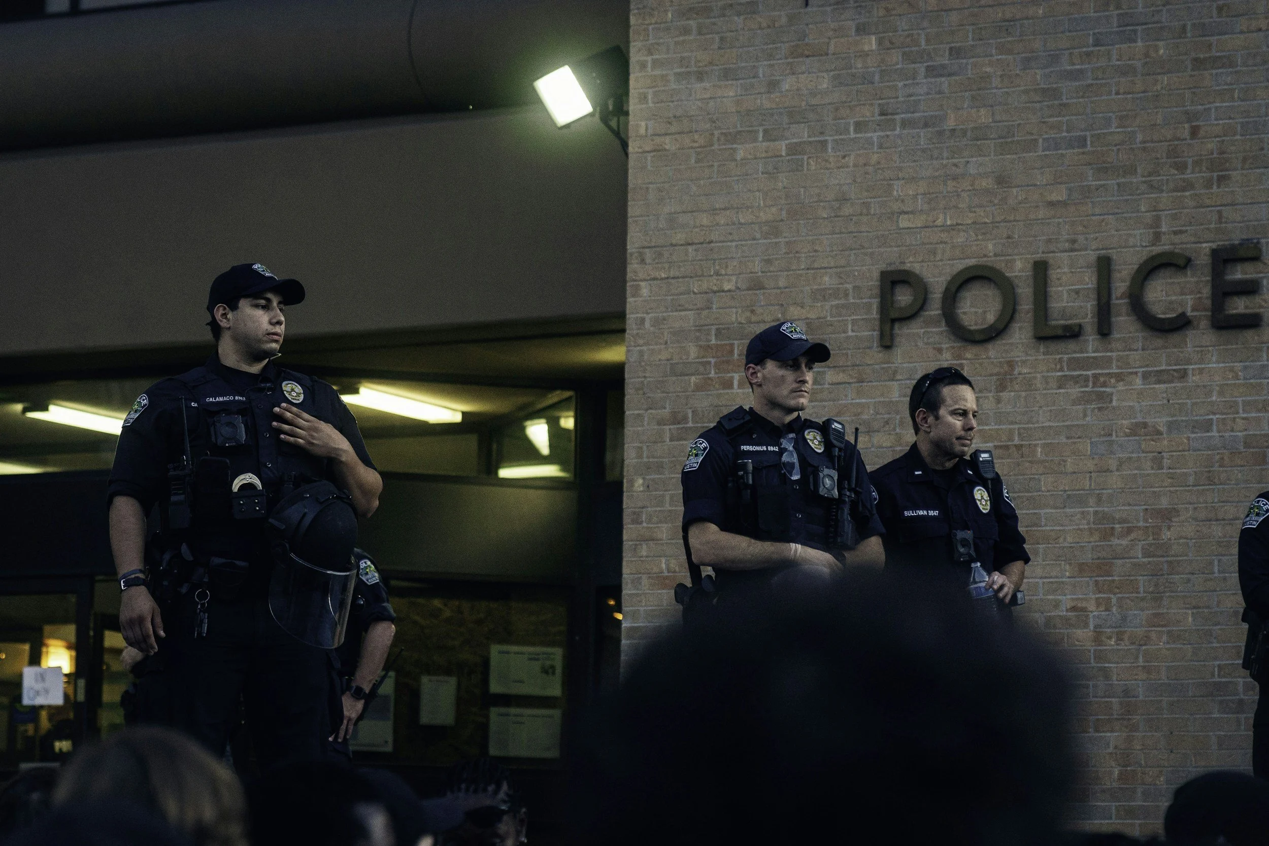 Three police officers standing against a brick wall outside a police station, with one officer placing his hand over his heart as others stand at attention.