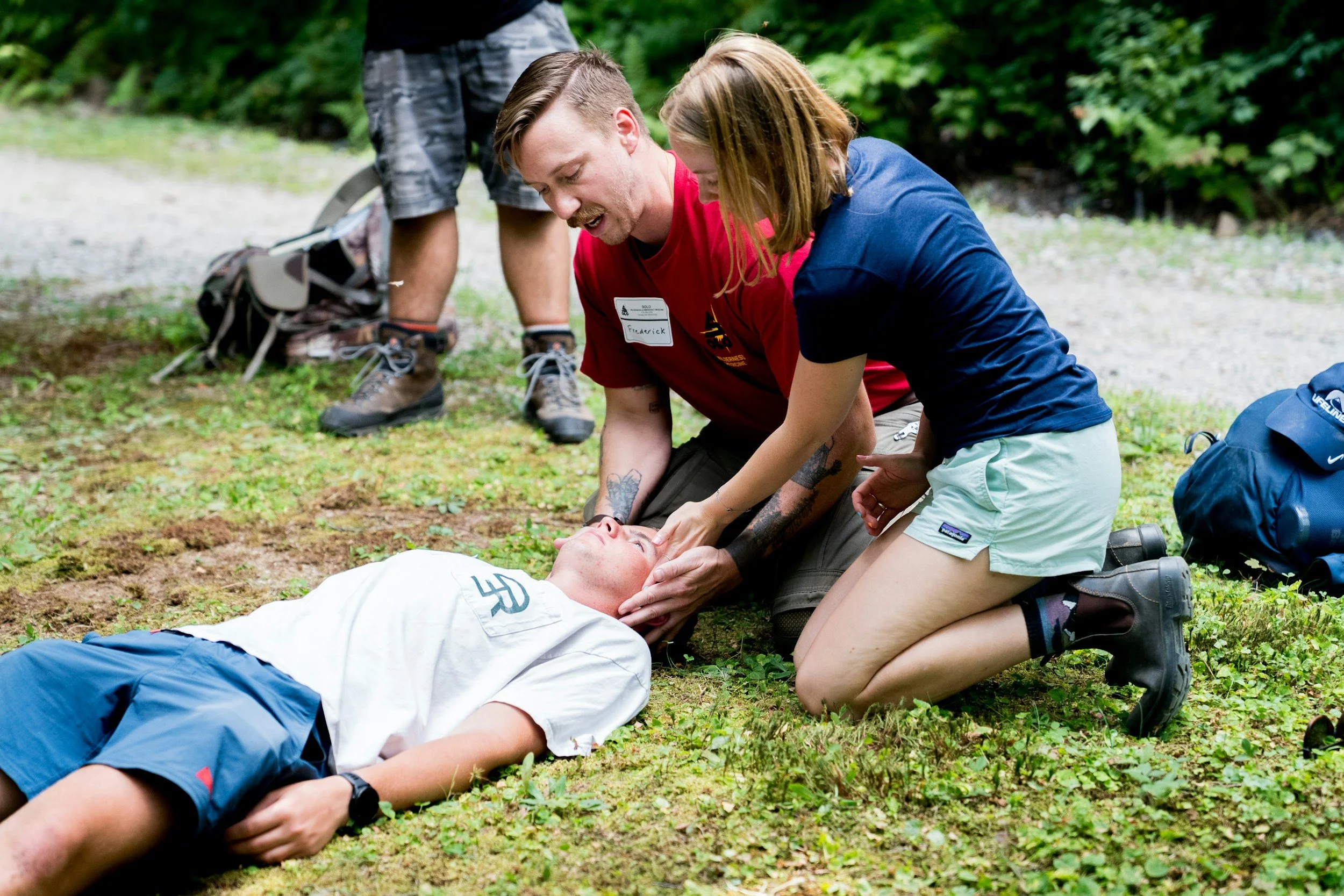 A person lies on the ground while others assist during a possible medical emergency in a forested outdoor setting.
