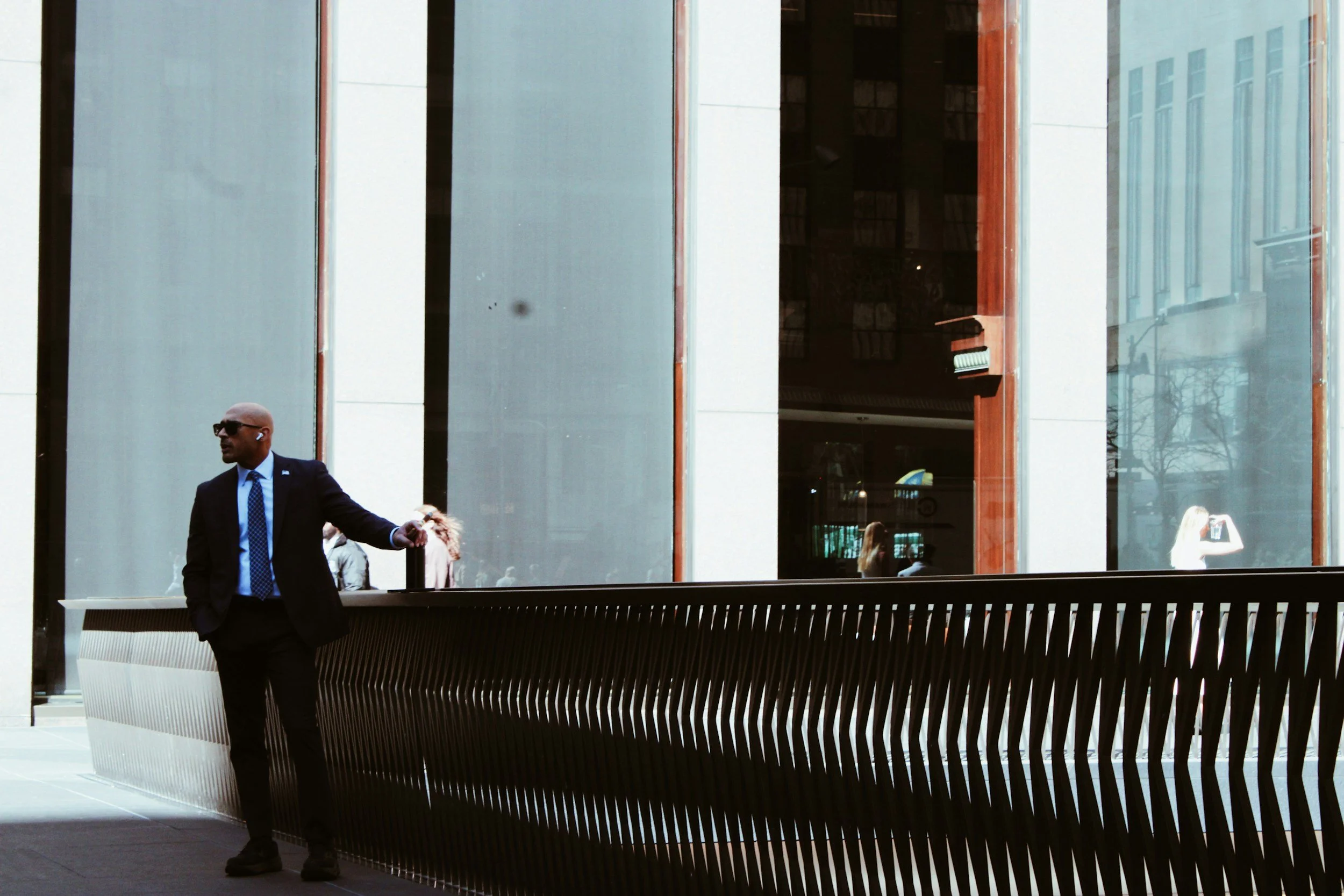 A man in a suit with sunglasses and earbuds standing outdoors in front of a modern building with large glass windows.